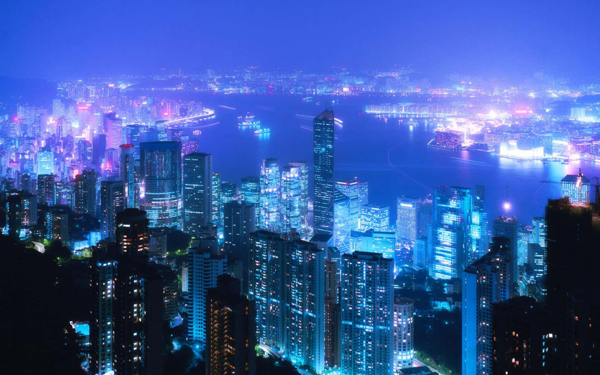 城市風景 桌布 壁紙 — 令人目眩的夜景，高聳的摩天大樓沿著充滿活力的海濱閃爍著藍色和紫色的燈光。