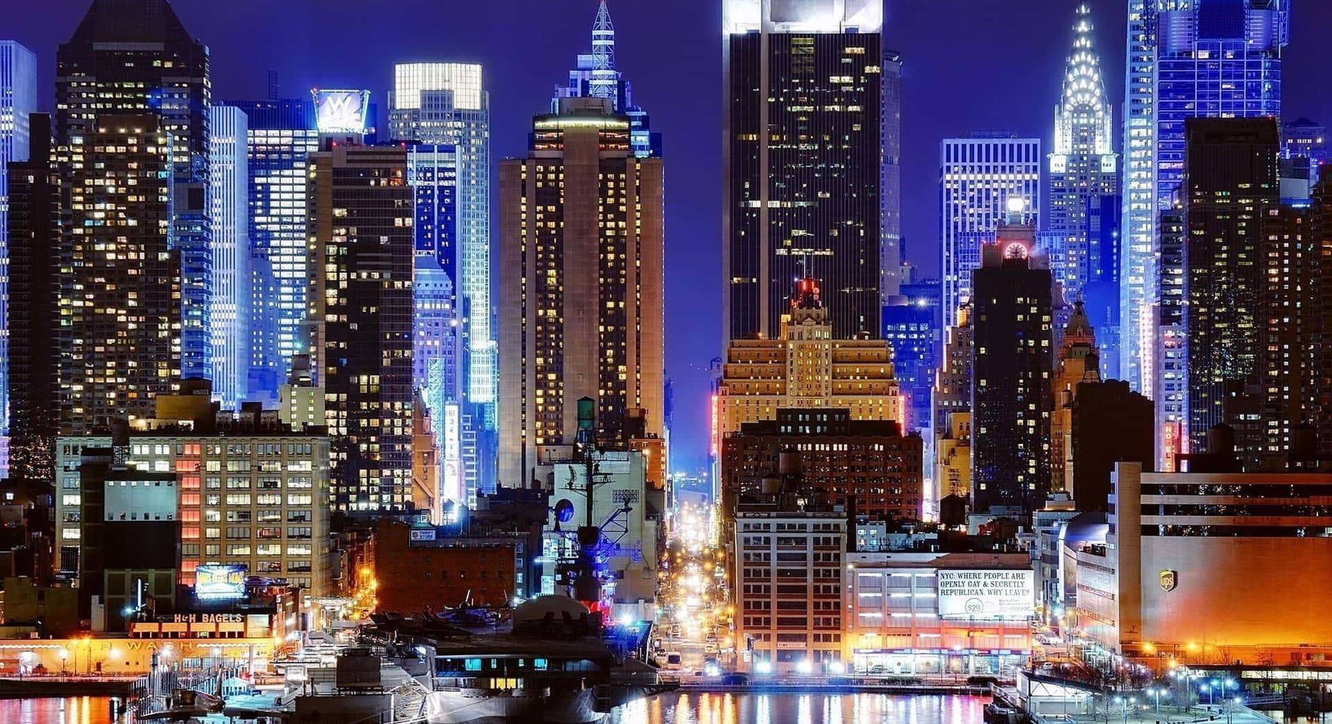 紐約市 桌布 壁紙 — 令人目眩的夜景城市景觀，高聳的摩天大樓閃爍著鮮豔的藍色、橙色和白色燈光，倒映在下方水面上。