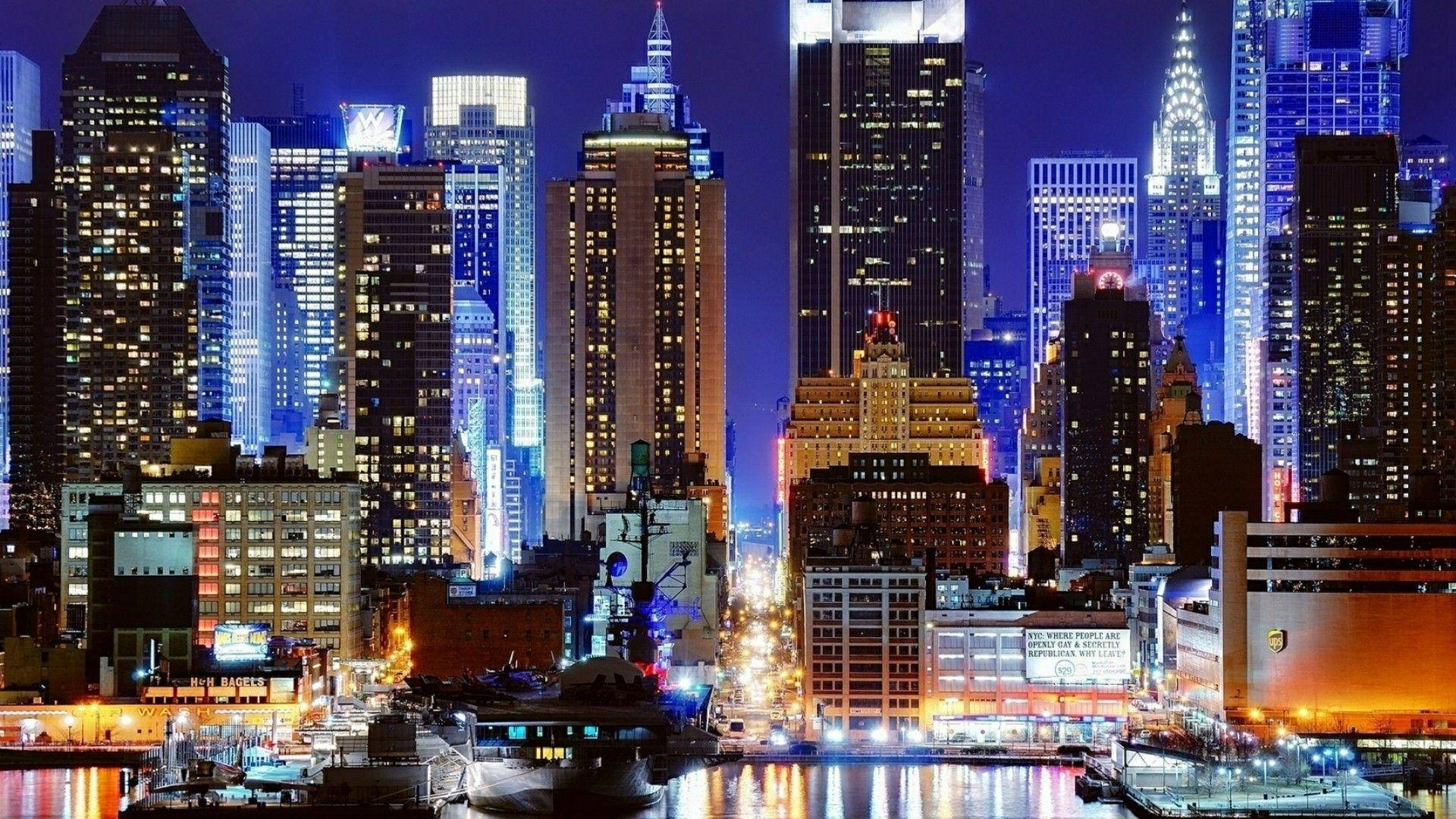 Skyscraper 桌布 壁紙 — 令人目眩的夜景城市景觀，高聳的摩天大樓閃爍著藍色和溫暖的黃色燈光，倒映在黑暗的水道上。