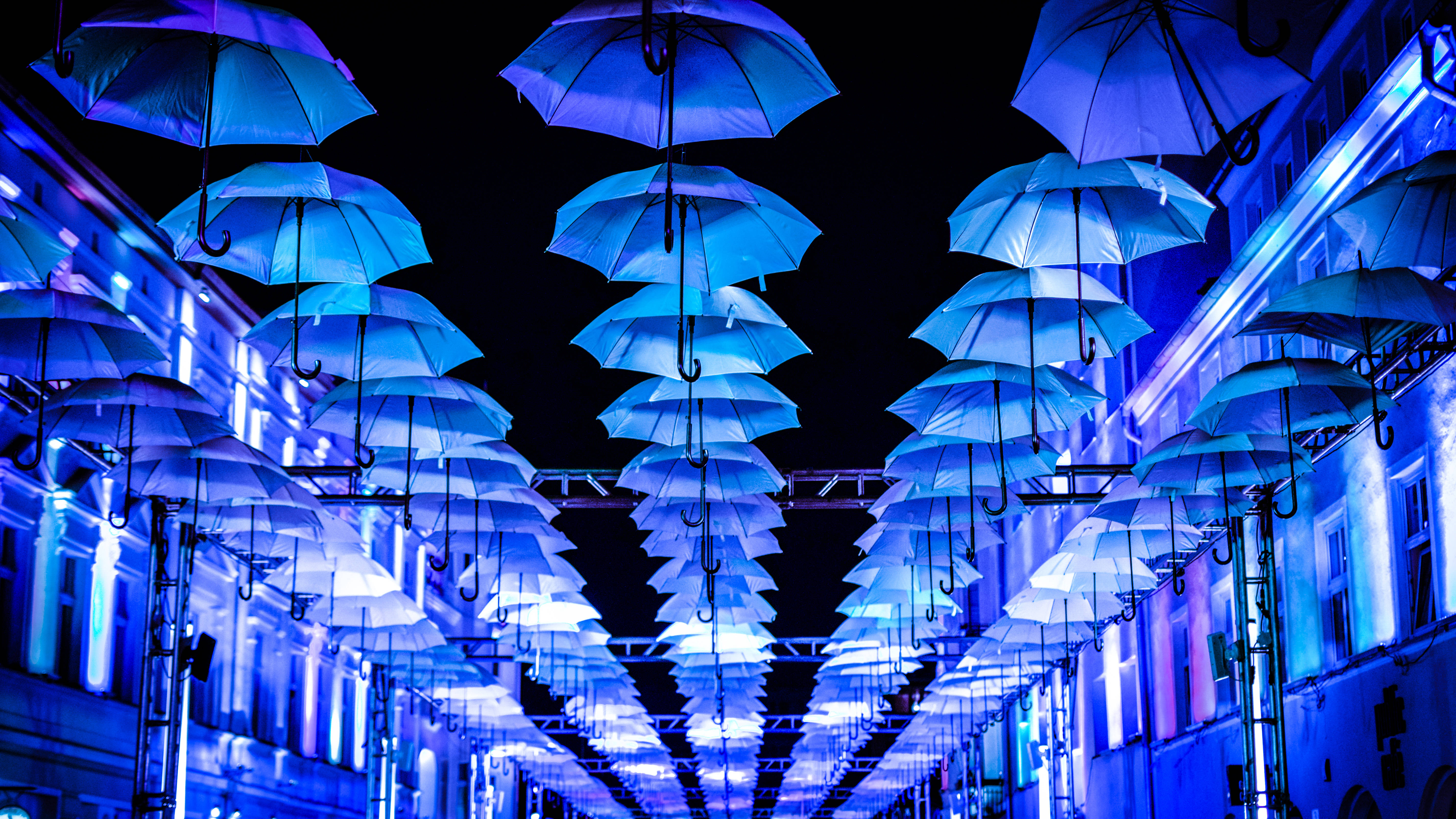 藍色 桌布 壁紙 — 令人眼花撩亂的夜景，發光的藍色雨傘懸掛在一條狹窄的城市街道上方，兩旁是燈火通明的建築物。