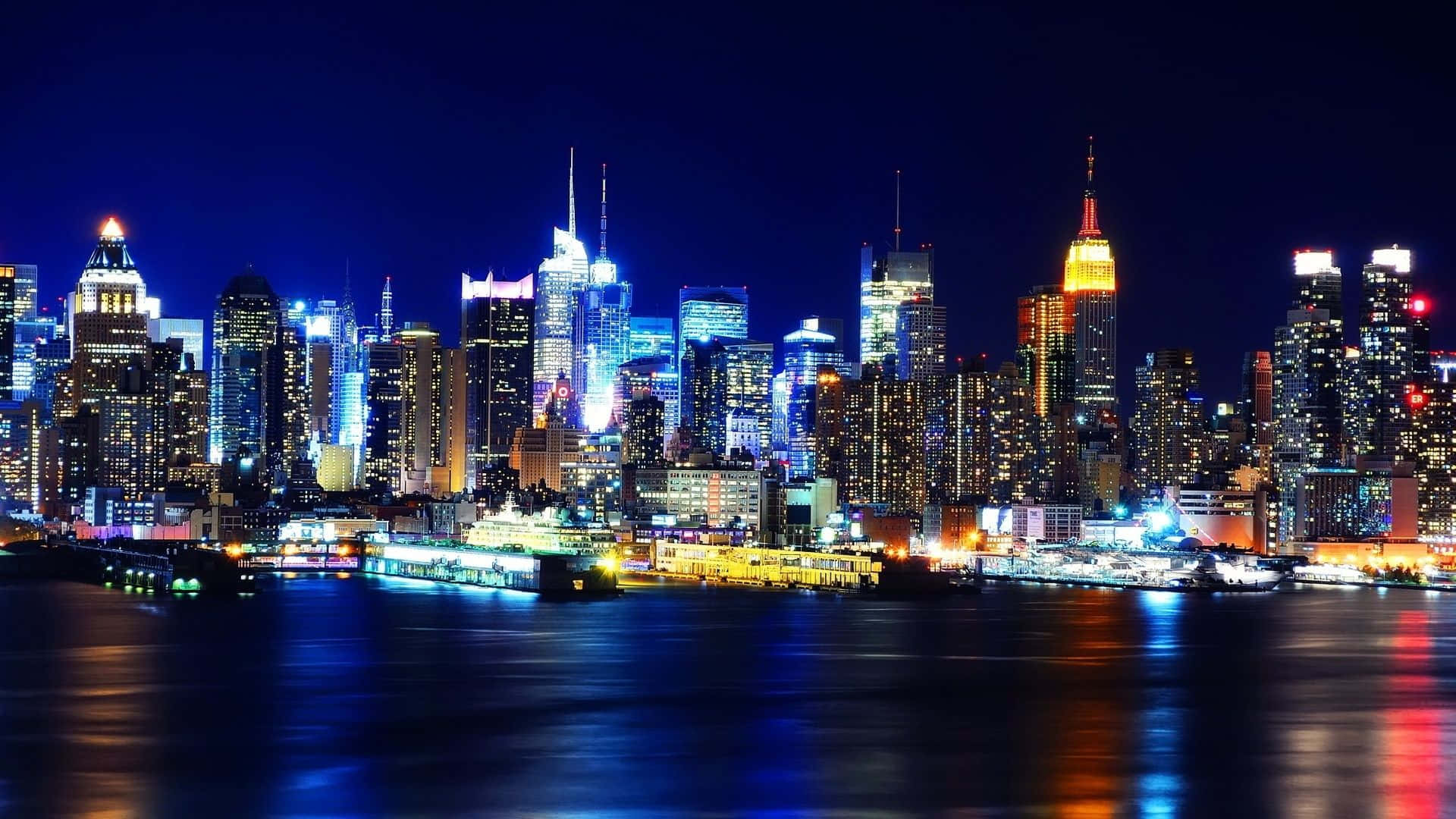 紐約市 桌布 壁紙 — 令人眼花撩亂的夜景，高聳的摩天大樓閃爍著藍色、黃色和白色的燈光，倒映在平靜的水面上。