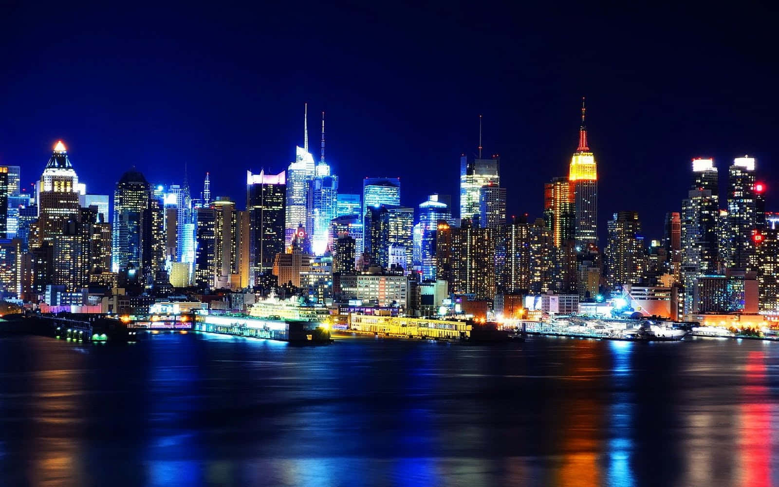 New York 桌布 壁紙 — 令人眼花撩亂的夜景，高聳的摩天大樓閃爍著霓虹燈，倒映在黑暗的水道上。