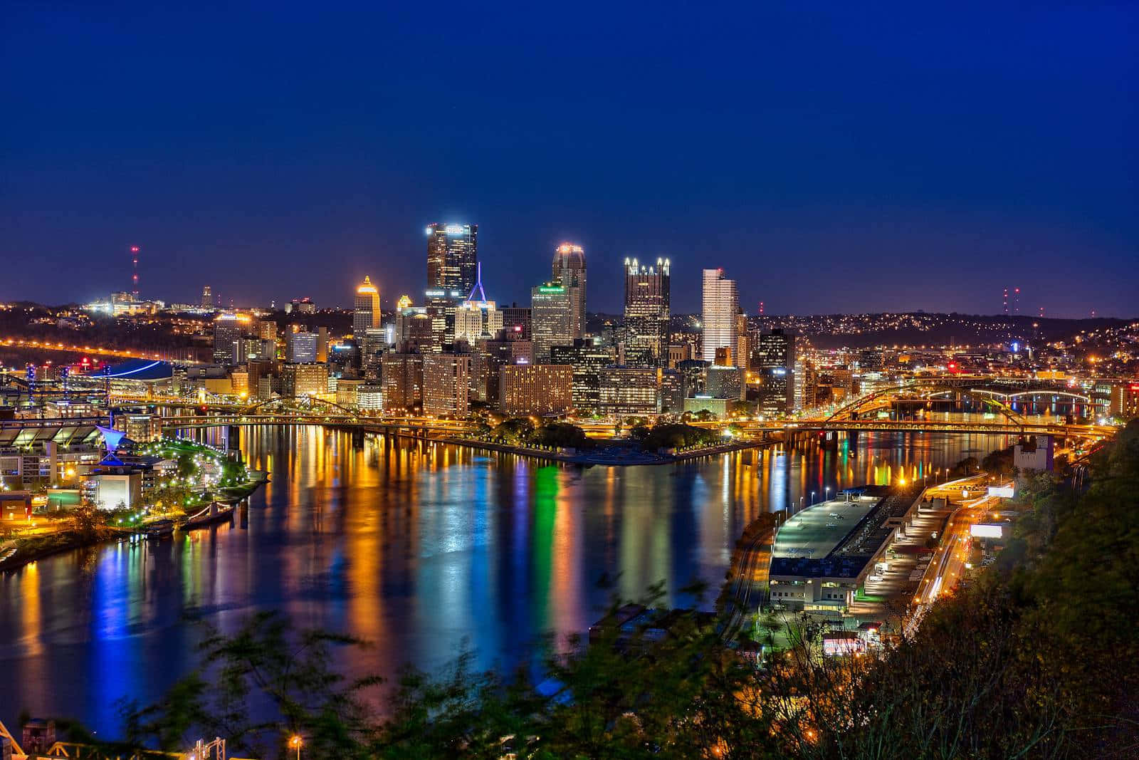 Pittsburgh Skyline 桌布 壁紙 — 令人眼花撩亂的夜間城市景觀，摩天大樓在深藍色天空下閃耀，燈光在下方平靜的河面上生動地倒映。