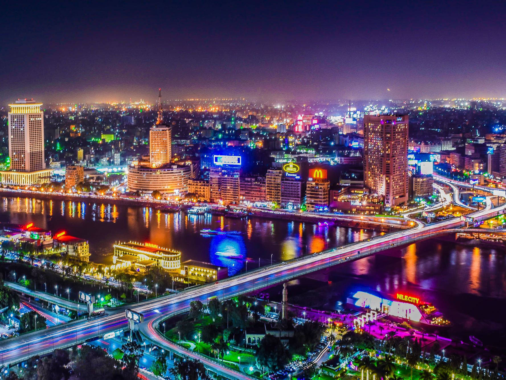 開羅 桌布 壁紙 — 令人眼花撩亂的夜間城市景觀，霓虹燈在河上反射，發光的橋樑連接充滿活力的城市區域。