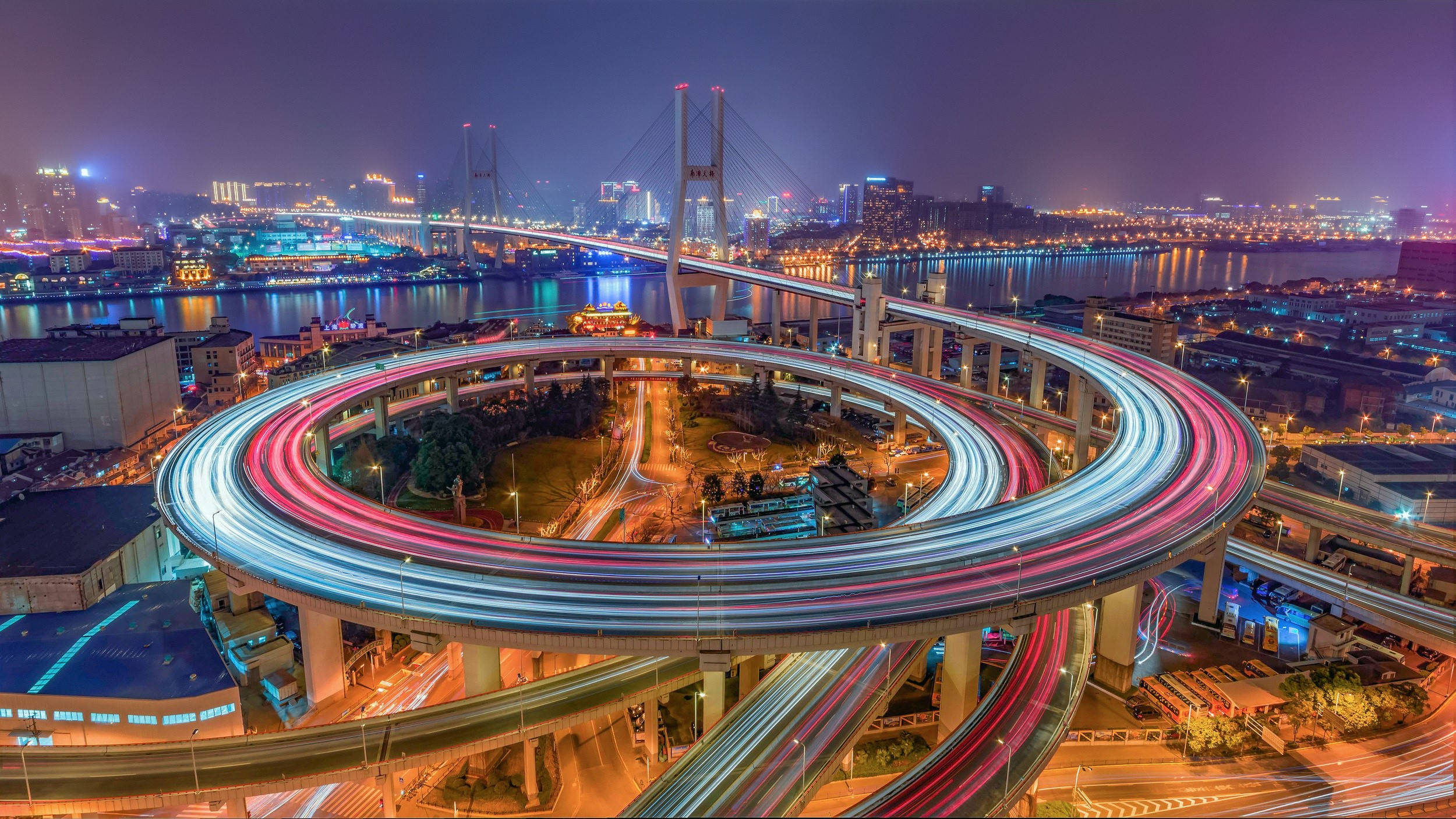 Asia 桌布 壁紙 — 令人眼花撩亂的夜間延時攝影，呈現螺旋形高速公路交匯處，車燈流光溢彩，照亮城市景觀和河流。