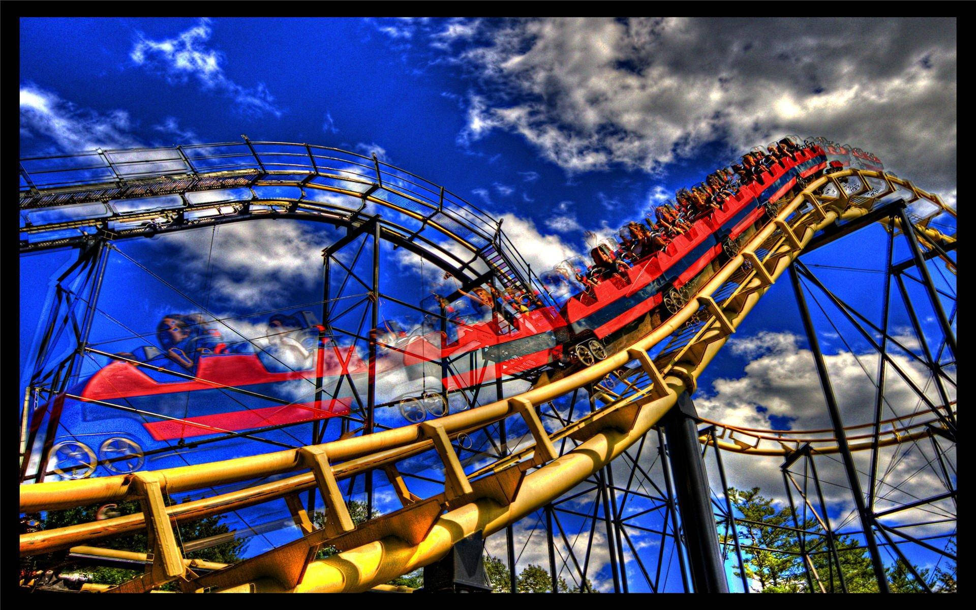 Theme Park 桌布 壁紙 — 令人興奮的紅色雲霄飛車，乘客尖叫著穿過蜿蜒的黃色軌道，背景是帶有戲劇性雲彩的鮮豔藍天。