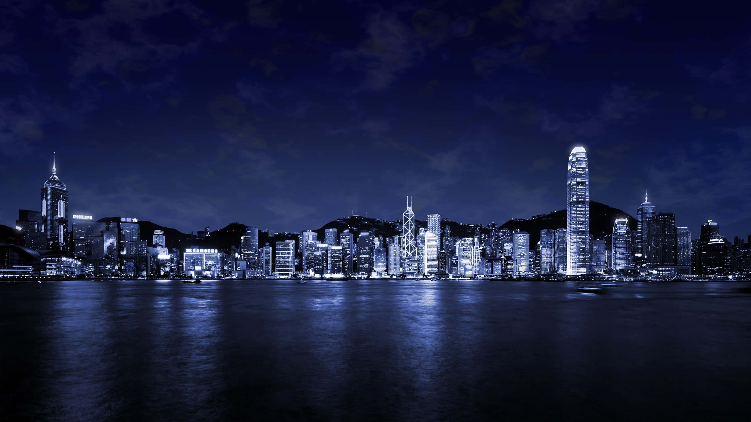 天際線 桌布 壁紙 — 令人驚嘆的單色香港夜景，摩天大樓在深藍色天空下閃爍，倒映在平靜的水面上。