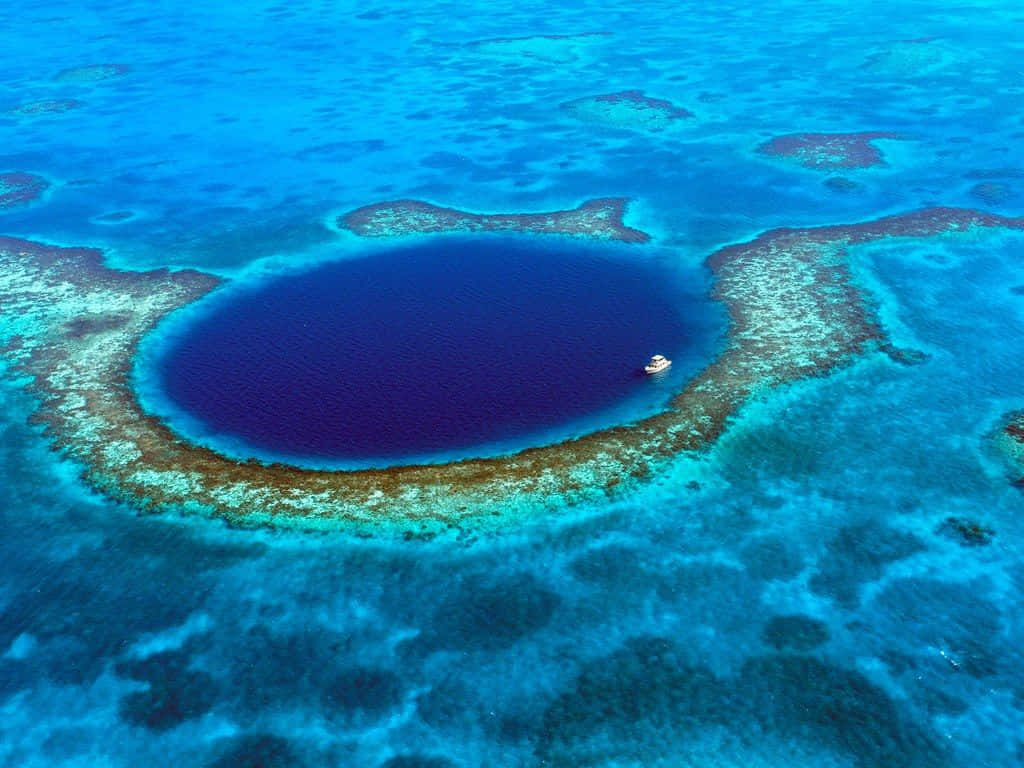 Blue Hole 桌布 壁紙 — 令人驚嘆的圓形藍色潟湖的鳥瞰圖，周圍環繞著充滿活力的珊瑚礁，中心漂浮著一艘小船。