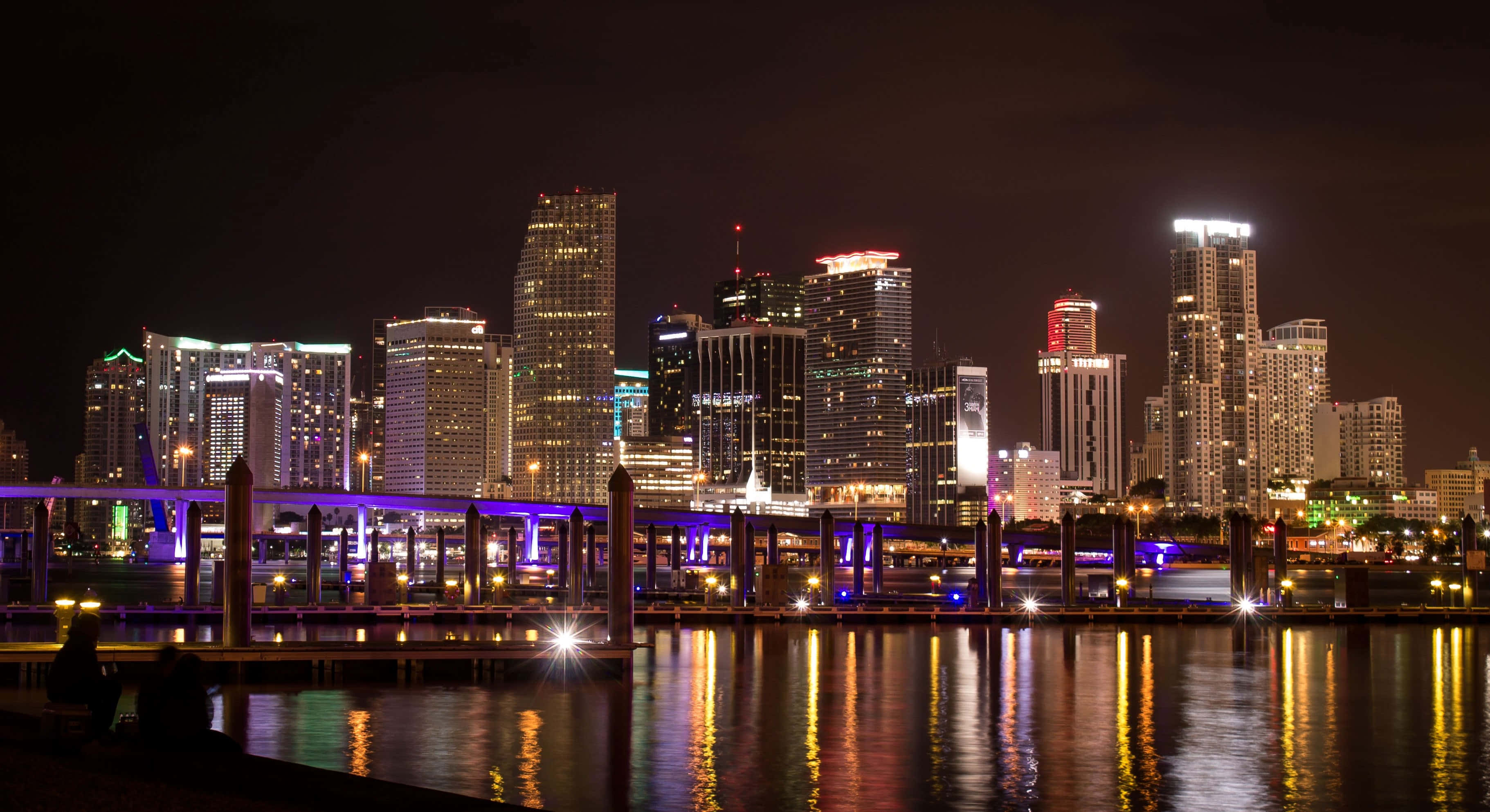 Miami 桌布 壁紙 — 令人驚嘆的夜景，摩天大樓在黑暗的天空中閃耀，它們的燈光倒映在下方平靜的水面上。