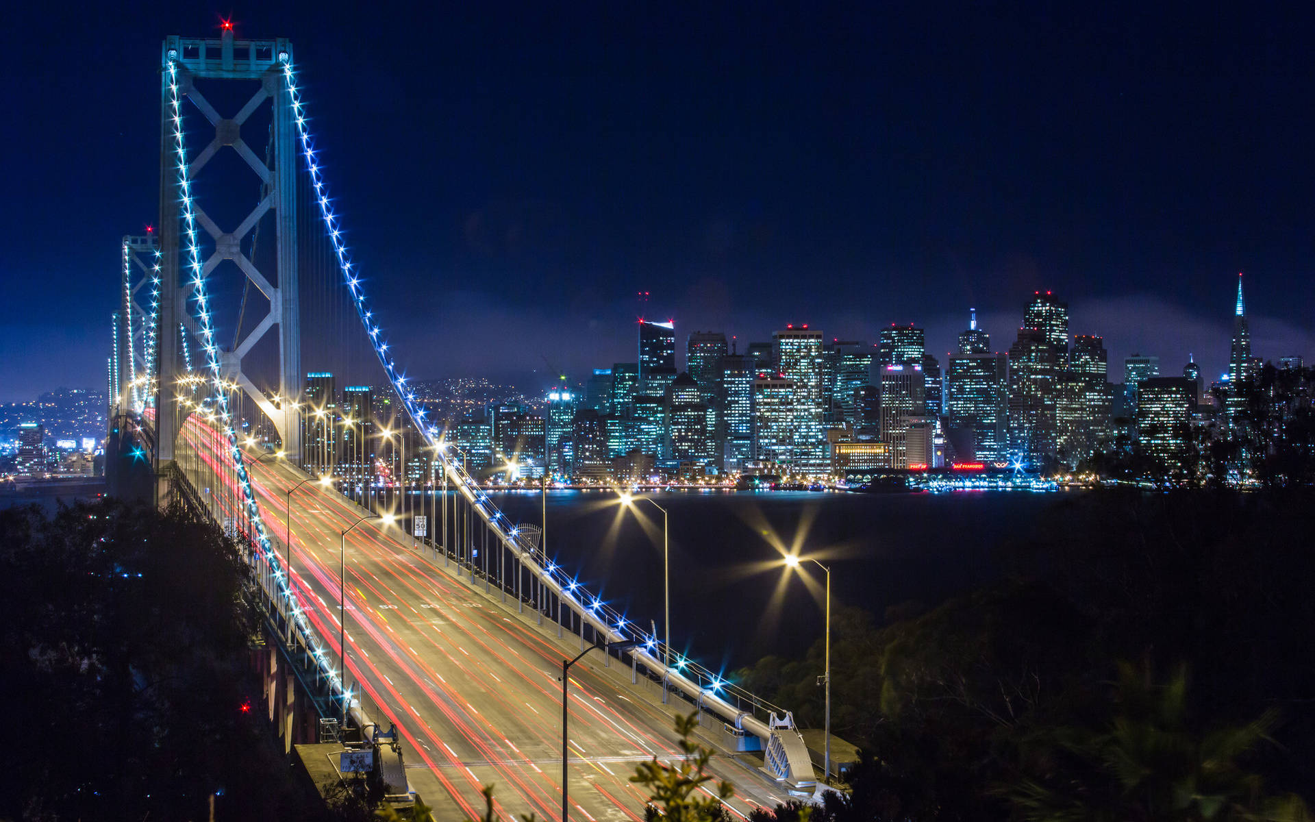 天際線 桌布 壁紙 — 令人驚嘆的夜景，照亮的海灣大橋，帶有流動的車燈和背景中閃爍的舊金山天際線。
