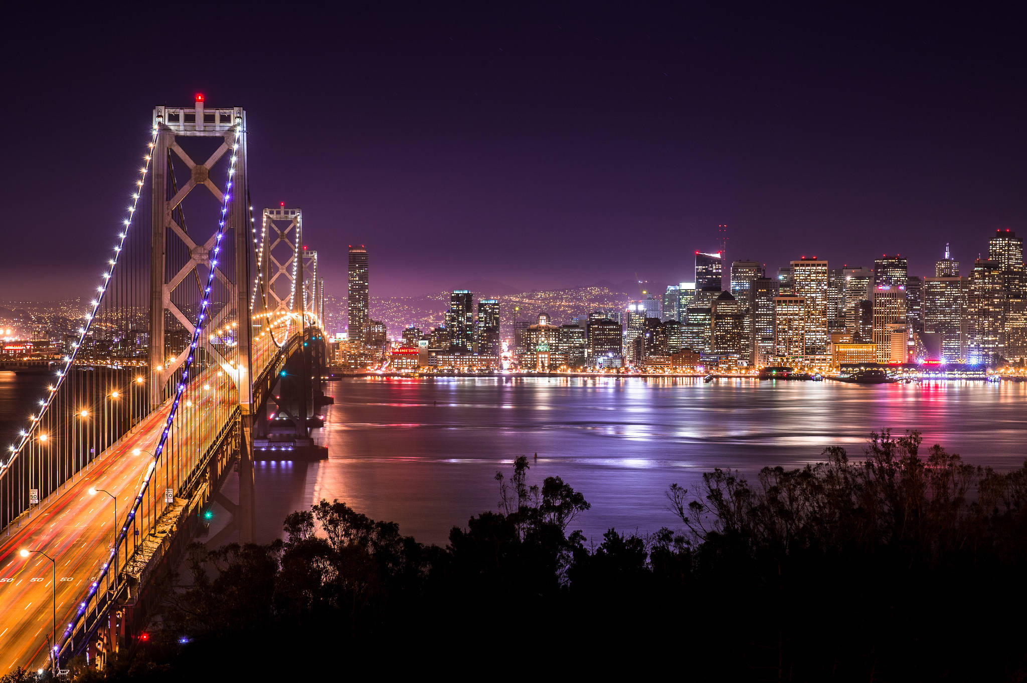 天際線 桌布 壁紙 — 令人驚嘆的夜景，照亮的海灣大橋和舊金山天際線倒映在平靜的水面上，前景有黑暗的樹木剪影。