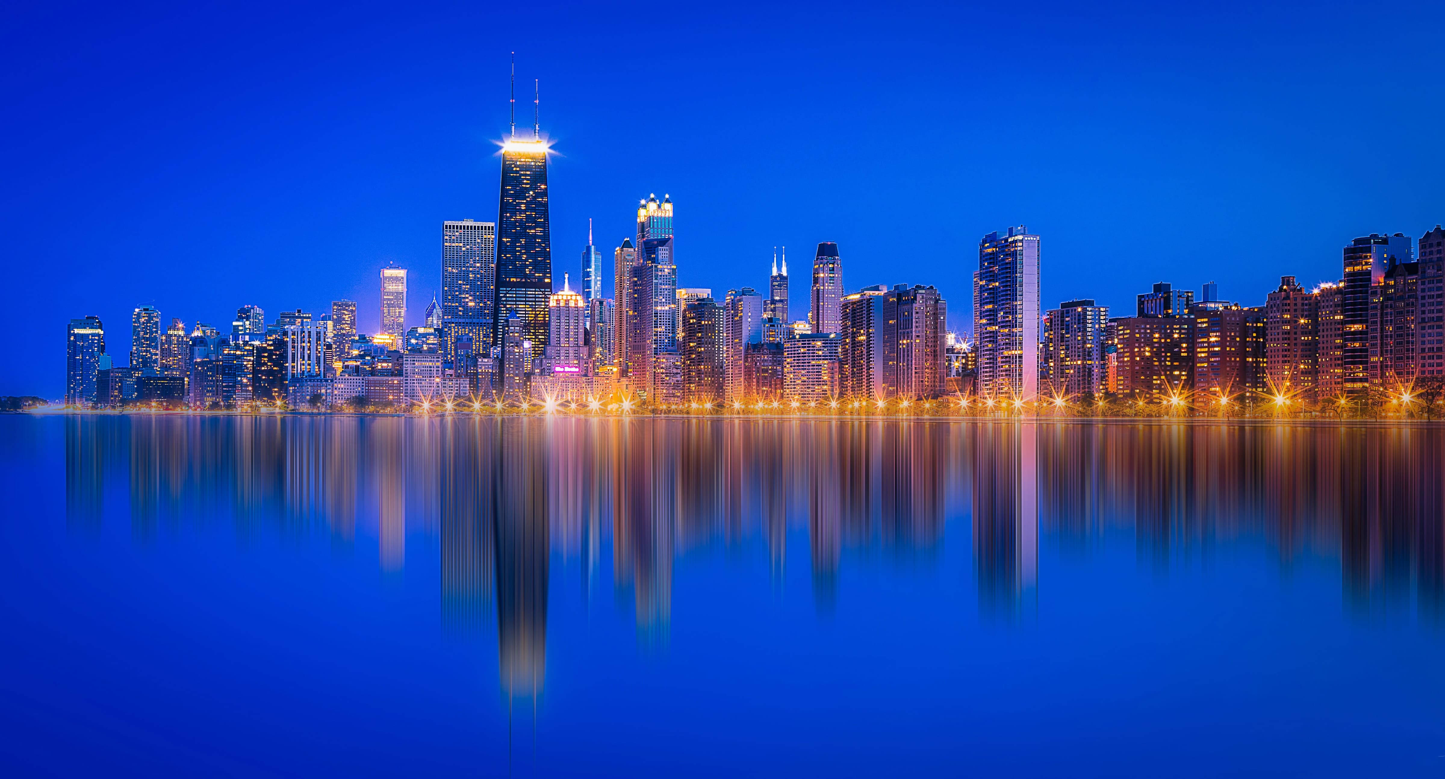 View 桌布 壁紙 — 令人驚嘆的夜景城市景觀，摩天大樓在深藍色天空下閃耀，完美倒映在下方平靜的水面上。