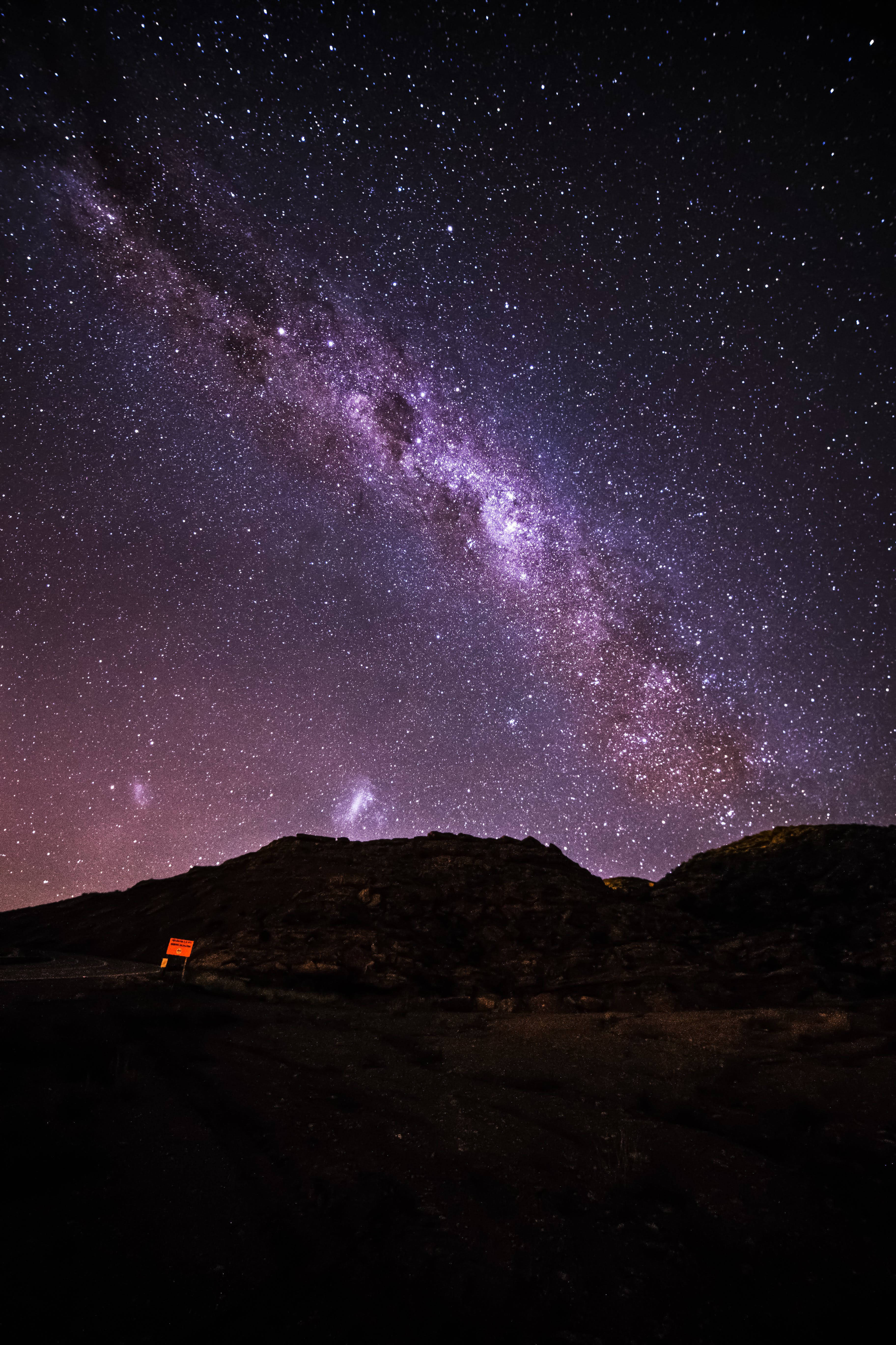 銀河 桌布 壁紙 — 令人驚嘆的夜空，佈滿繁星，充滿活力的紫色銀河在黑暗崎嶇的山丘上拱起，下方有一個孤獨的發光標誌。