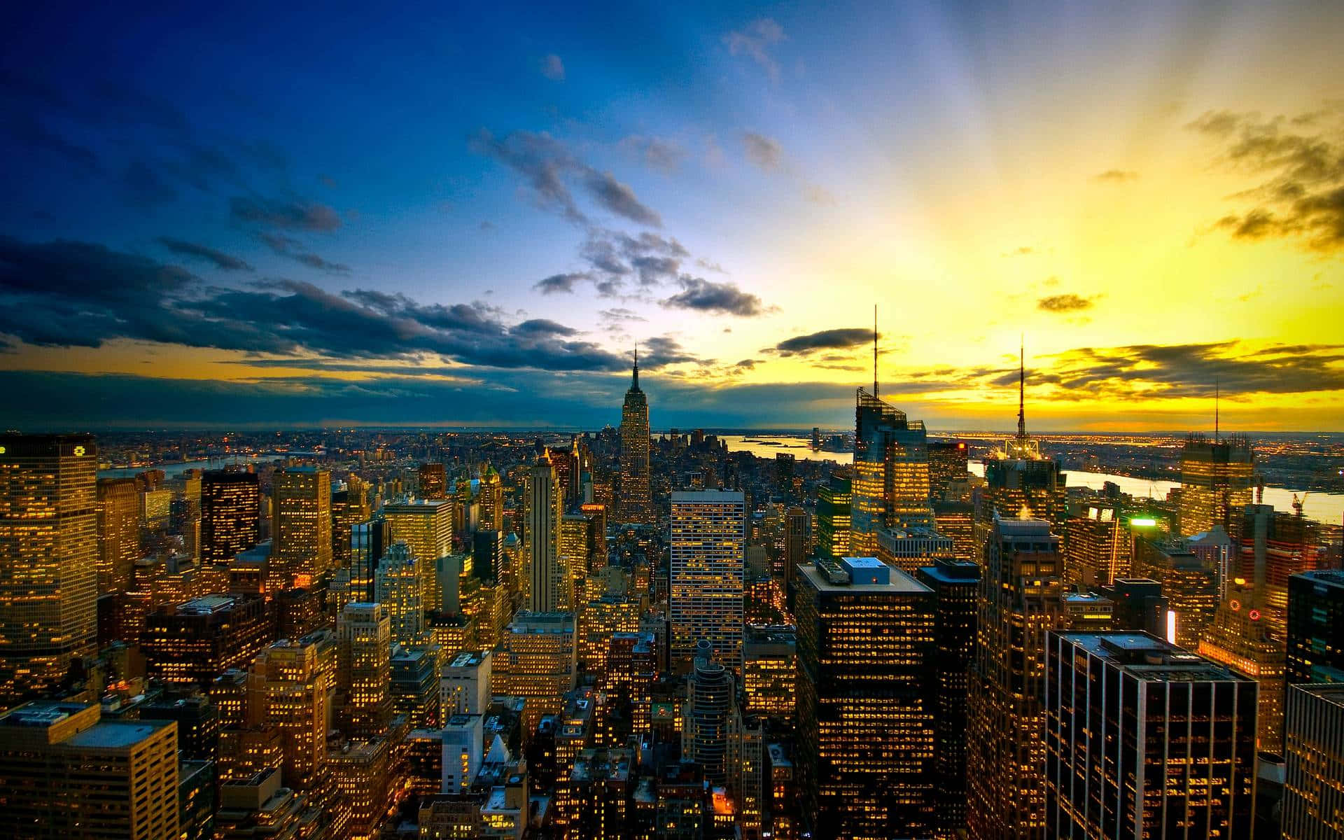 New York 桌布 壁紙 — 令人驚嘆的日落，映照在密集的城市天際線上，金色光芒從摩天大樓後方放射而出，下方城市燈光閃爍。