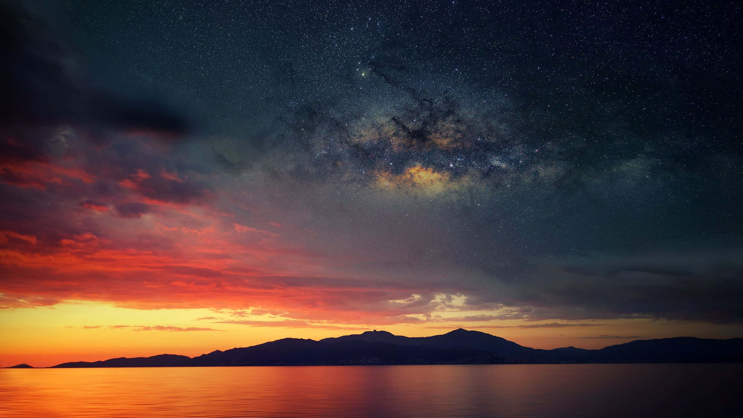 Ocean Sunset 桌布 壁紙 — 令人驚嘆的暮色天空，火橙色雲朵與繁星點點的銀河相遇，銀河拱跨過剪影般的山脈和寧靜的水面。