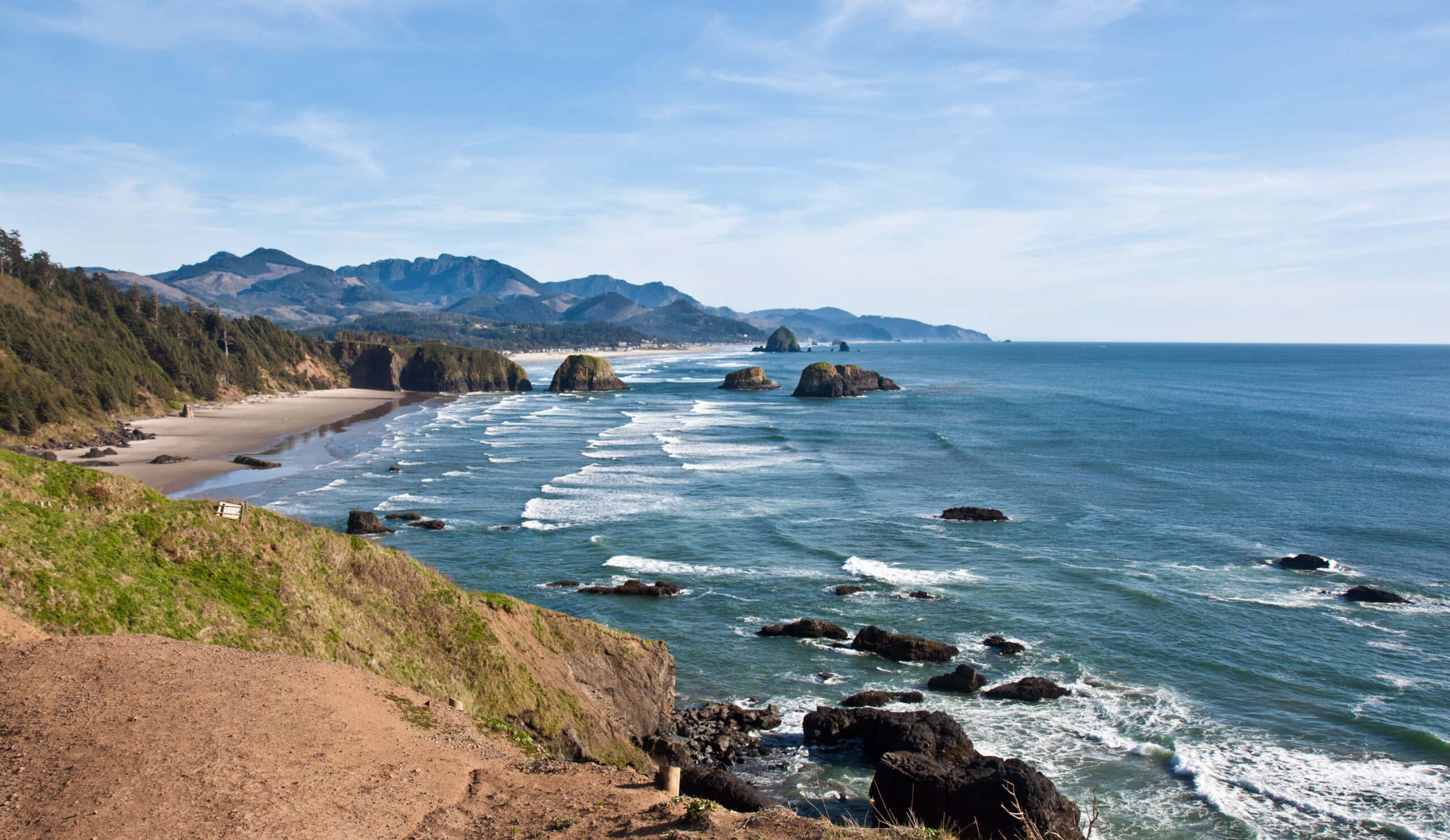 Oregon Coast 桌布 壁紙 — 令人驚嘆的海岸景觀，碧綠色的海浪拍打著深色岩石，沙灘坐落在綠色懸崖和遠處山脈之間，天空清澈湛藍。