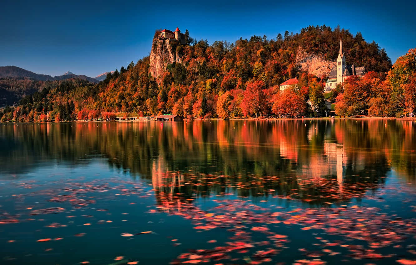 Slovenia 桌布 壁紙 — 令人驚嘆的秋日湖邊景色，懸崖上的城堡和教堂坐落在火紅、橙色和金色的樹木中，倒映在平靜的水面上。