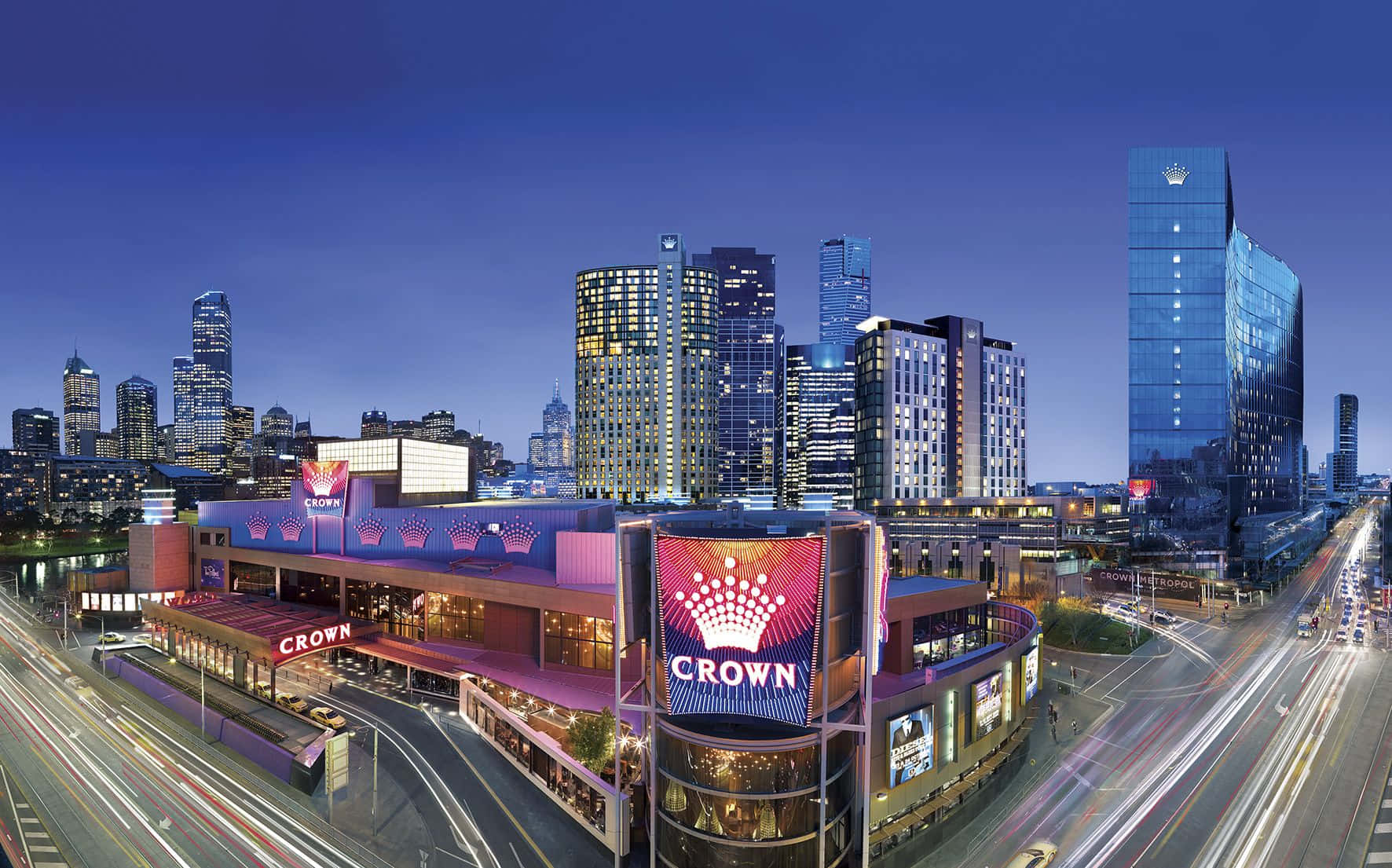 Road 桌布 壁紙 — 令人驚嘆的黃昏城市景觀， featuring Crown 賭場，閃爍的燈光和車流劃過深藍色的天空。