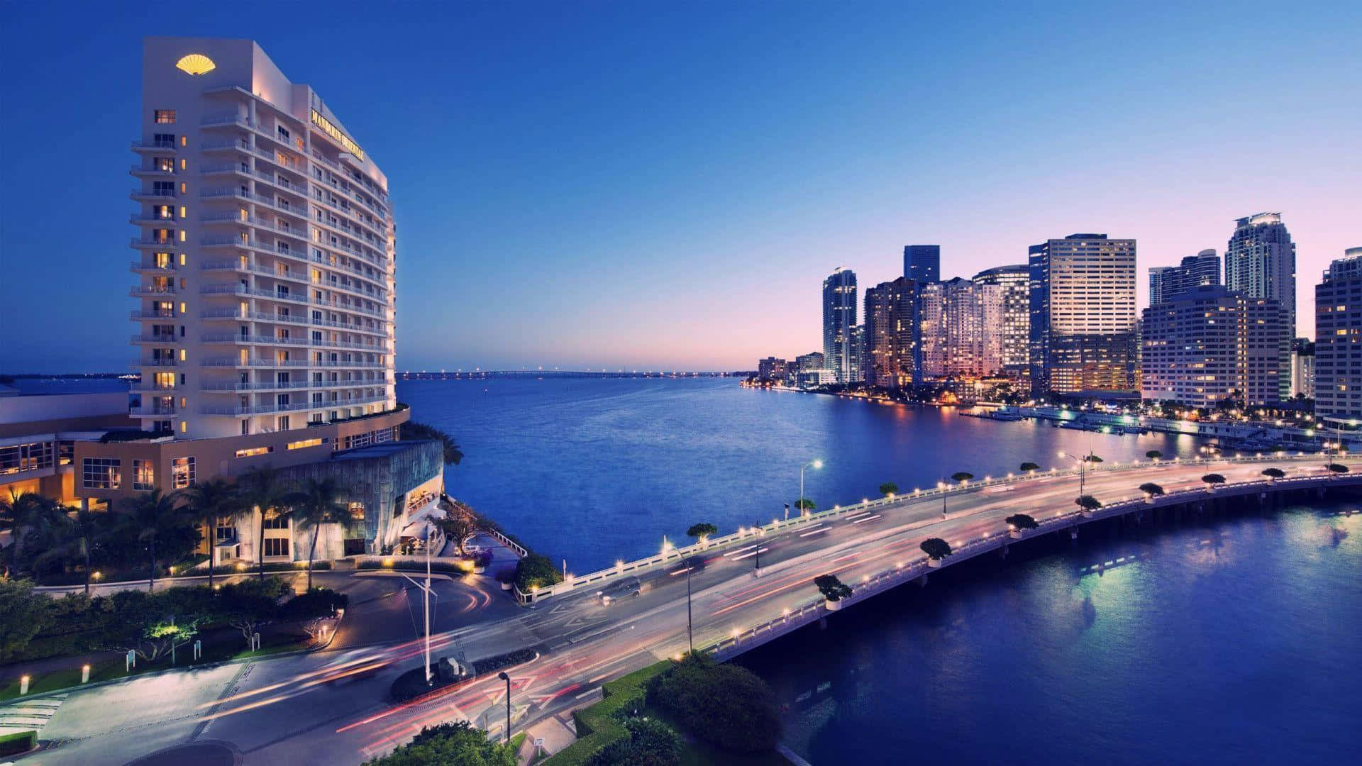 Miami 桌布 壁紙 — 令人驚嘆的黃昏城市景觀，發光的酒店和橋樑橫跨水面，在深藍色天空的映襯下被溫暖的燈光照亮。