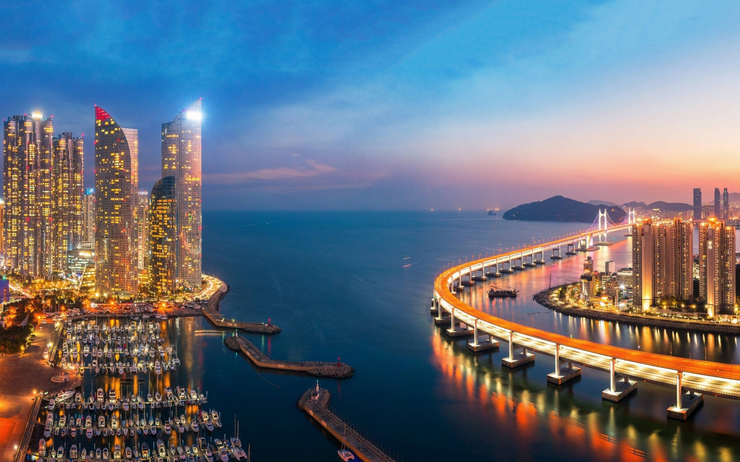 Busan 桌布 壁紙 — 令人驚嘆的黃昏城市景觀，閃爍的摩天大樓、彎曲的照明橋樑和停滿船隻的碼頭，倒映在平靜的水面上。