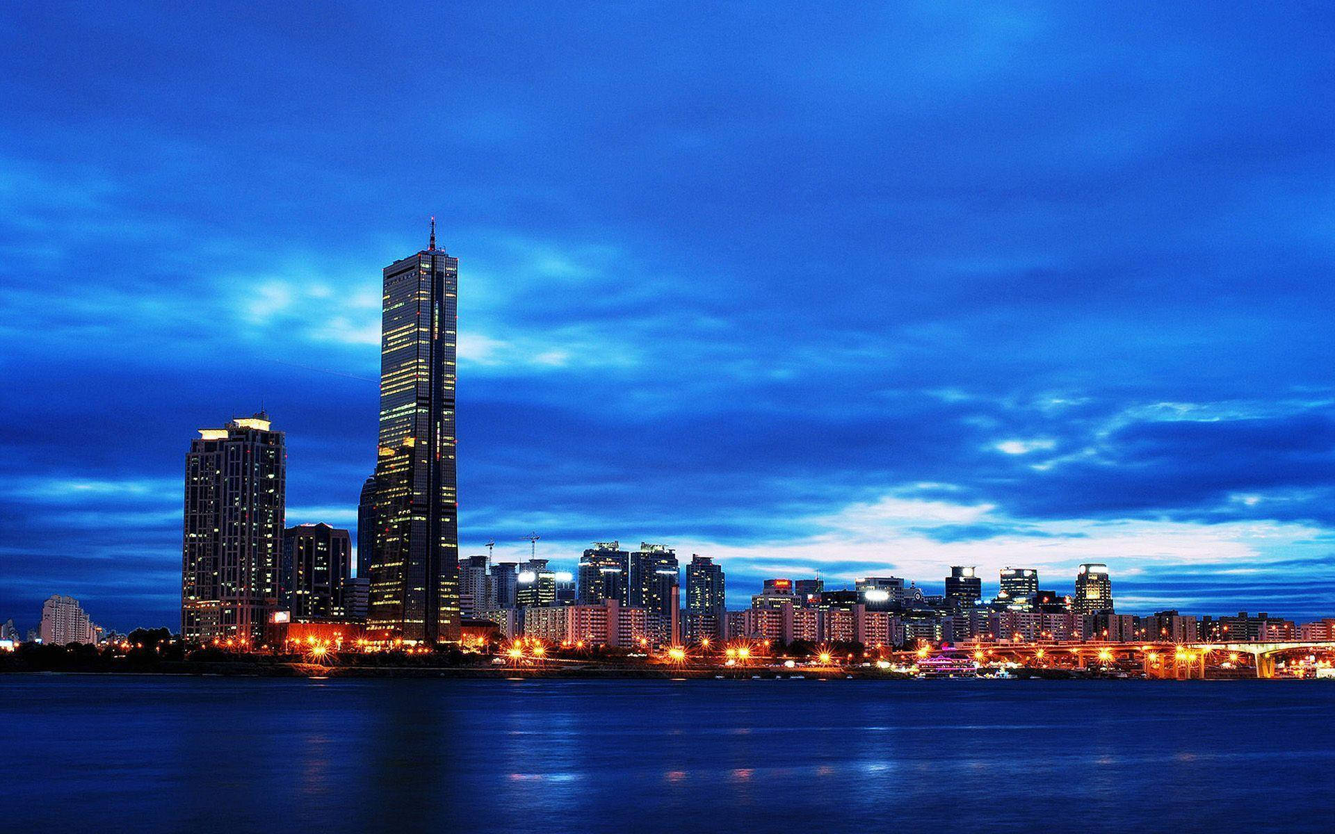 南韓 桌布 壁紙 — 令人驚嘆的黃昏城市景觀，高聳的摩天大樓在深藍色天空下，將溫暖的燈光反射在平靜的水面上。