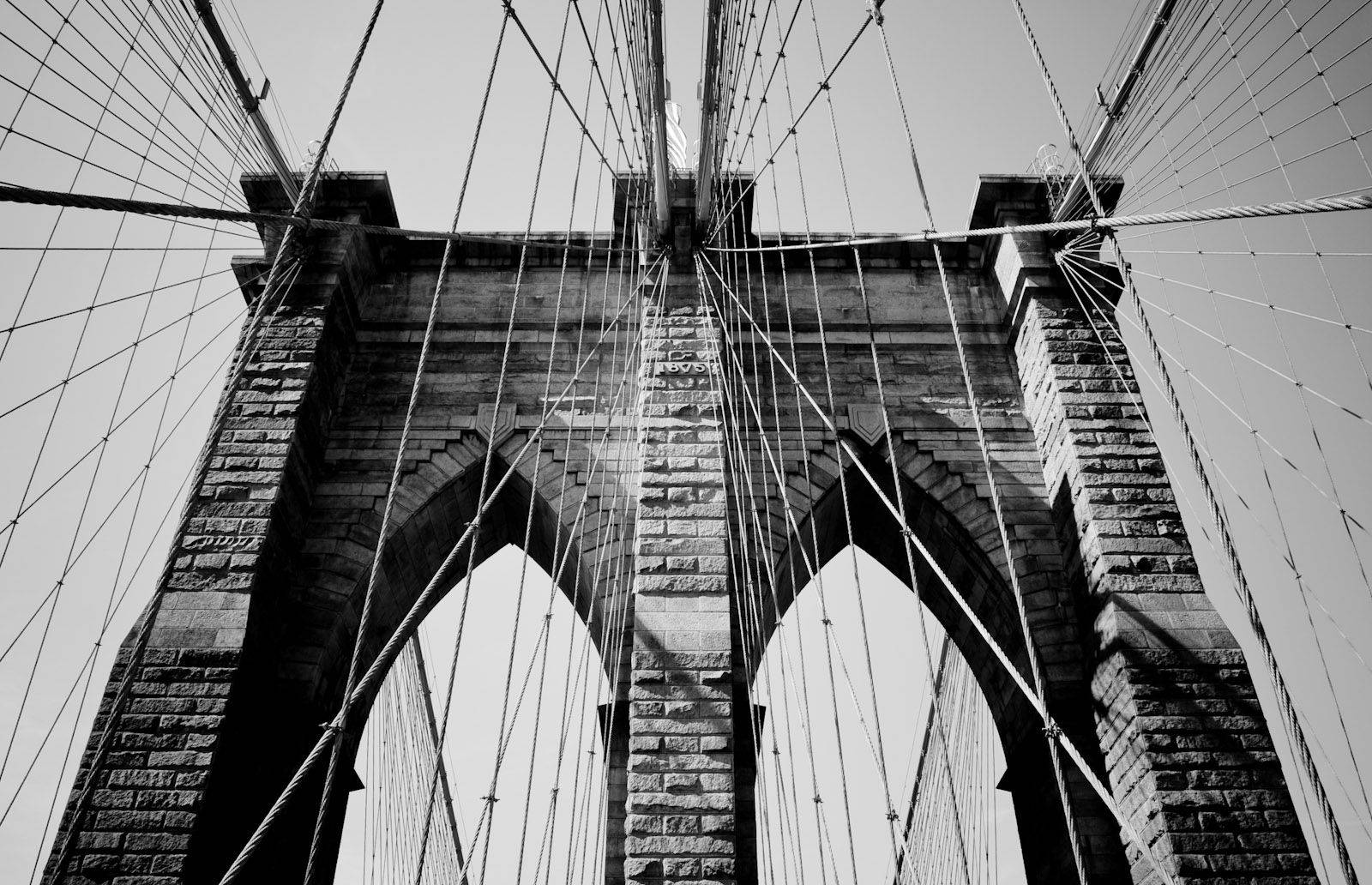 New York 桌布 壁紙 — 以低角度拍攝布魯克林大橋的石拱和懸索纜線，襯托著晴朗天空的戲劇性黑白照片。