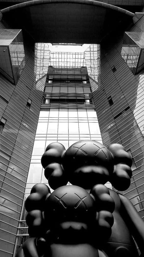 kaws pc 桌布 壁紙 — 以低角度拍攝的單色照片，巨型KAWS雕塑被高聳的玻璃和鋼鐵摩天大樓框住。