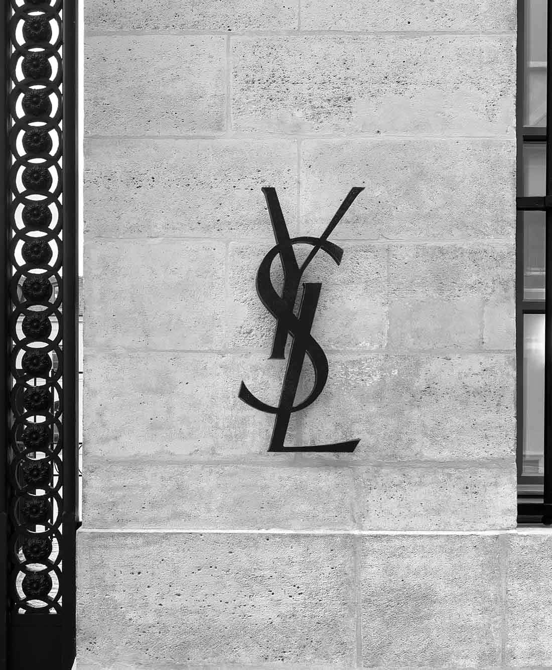 YSL 桌布 壁紙 — 以單色特寫拍攝的標誌性YSL標誌，安裝在帶有紋理的石牆上，兩側飾有華麗的黑色鐵藝。