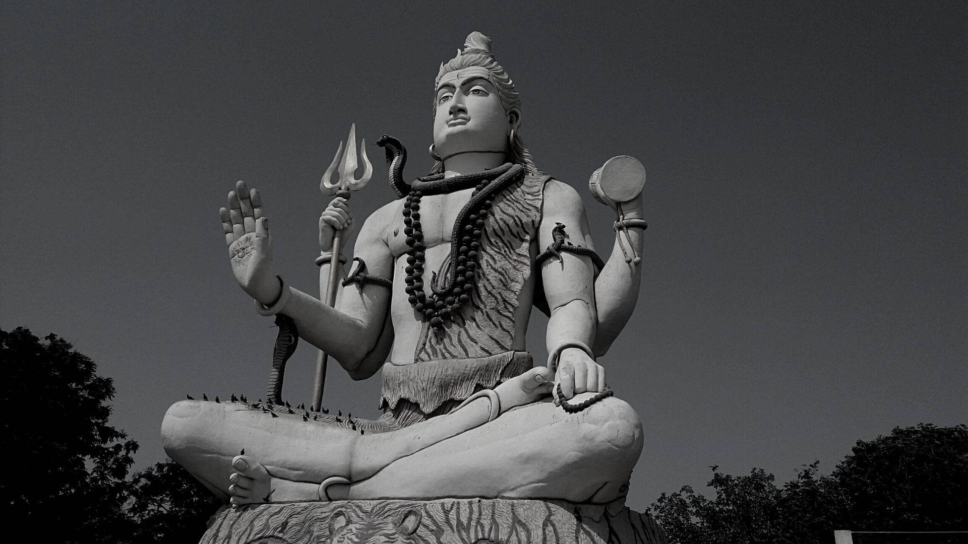shiva dark 桌布 壁紙 — 以多雲天空為背景，濕婆神手持三叉戟和鼓的冥想姿勢的紀念性黑白雕像。