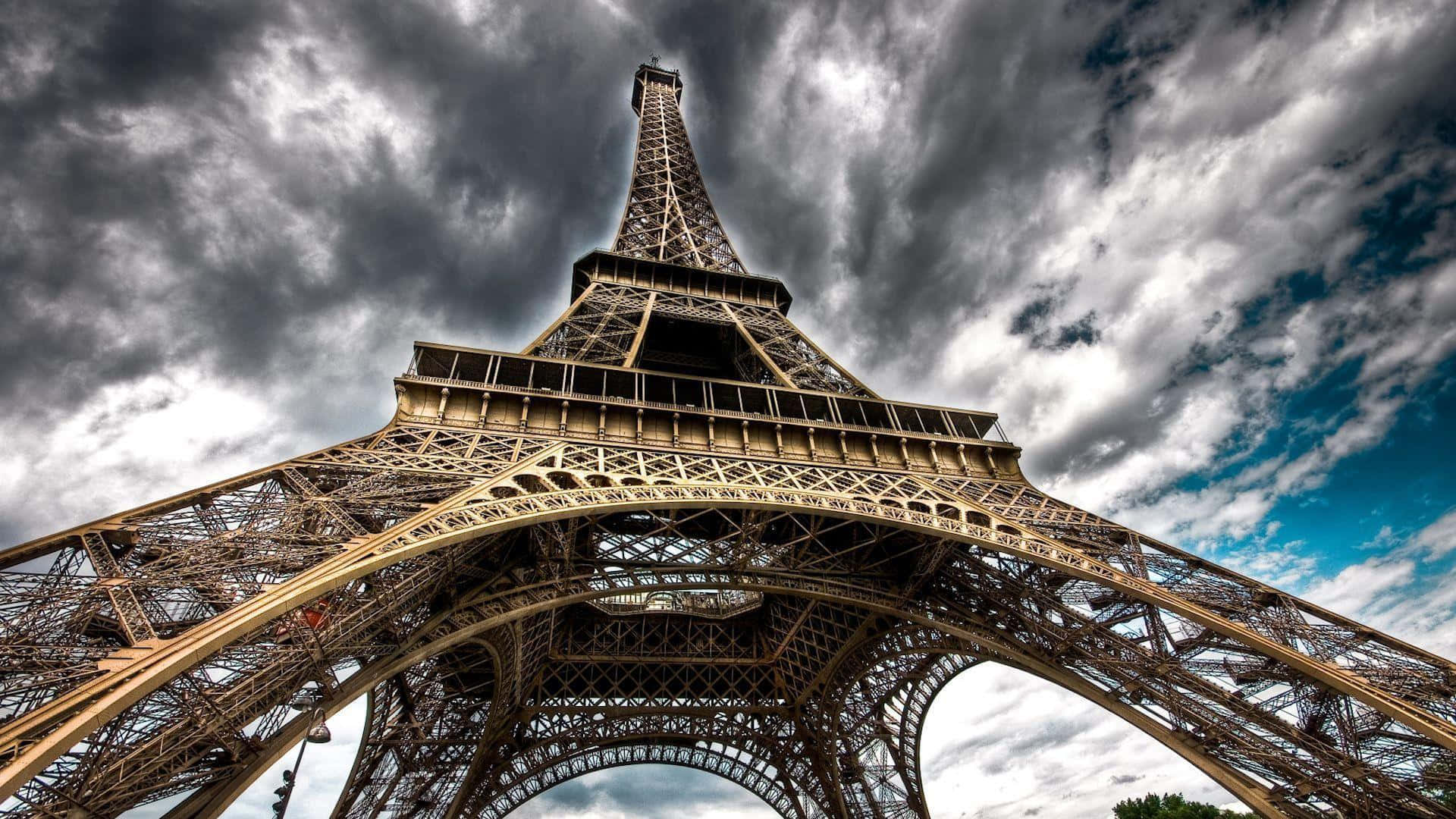 Eiffel Tower 桌布 壁紙 — 以帶有藍色斑塊的戲劇性多雲天空為背景，從低角度觀看艾菲爾鐵塔複雜的鐵格子結構。