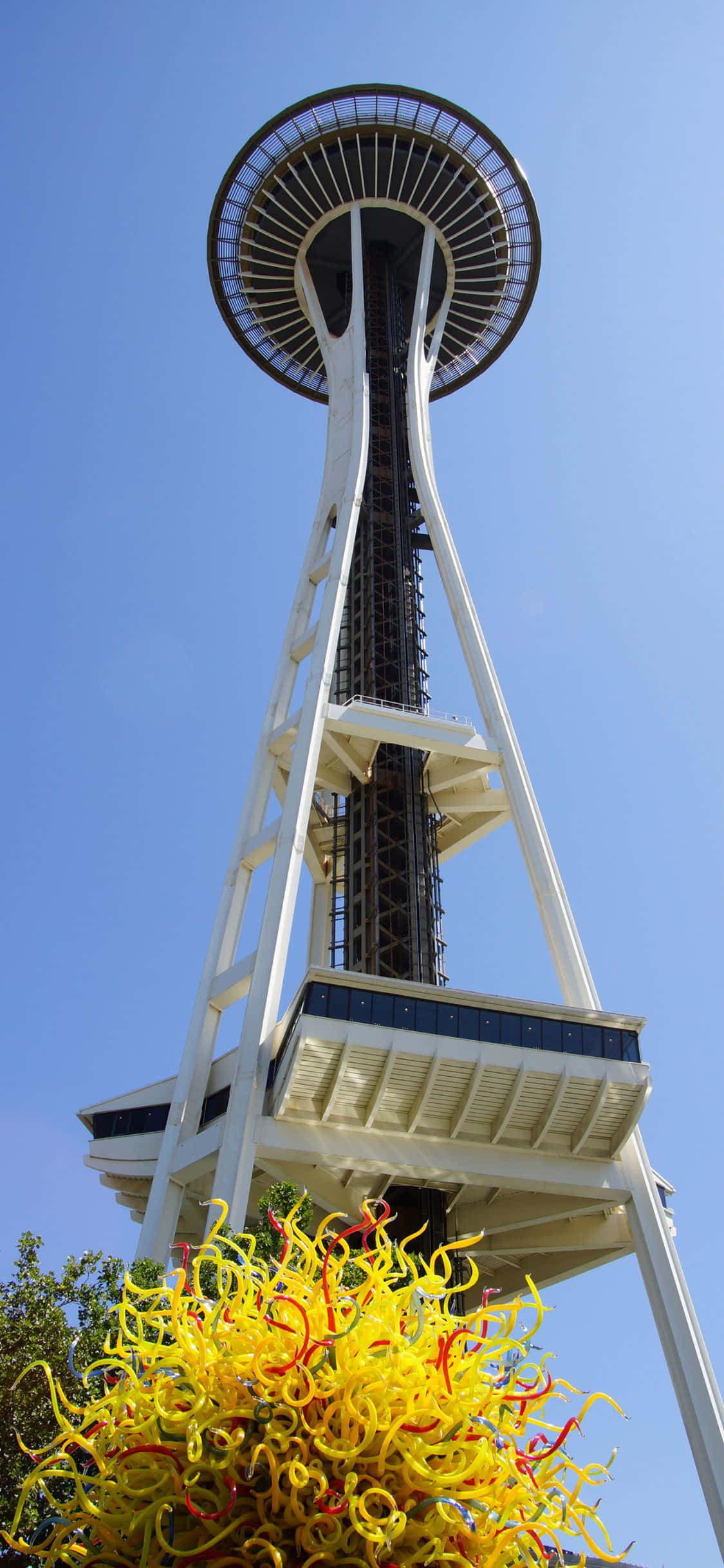 iPhone X Seattle 桌布 壁紙 — 以清晰藍天為背景的低角度拍攝太空針塔，前景有一座充滿活力的黃色玻璃雕塑。