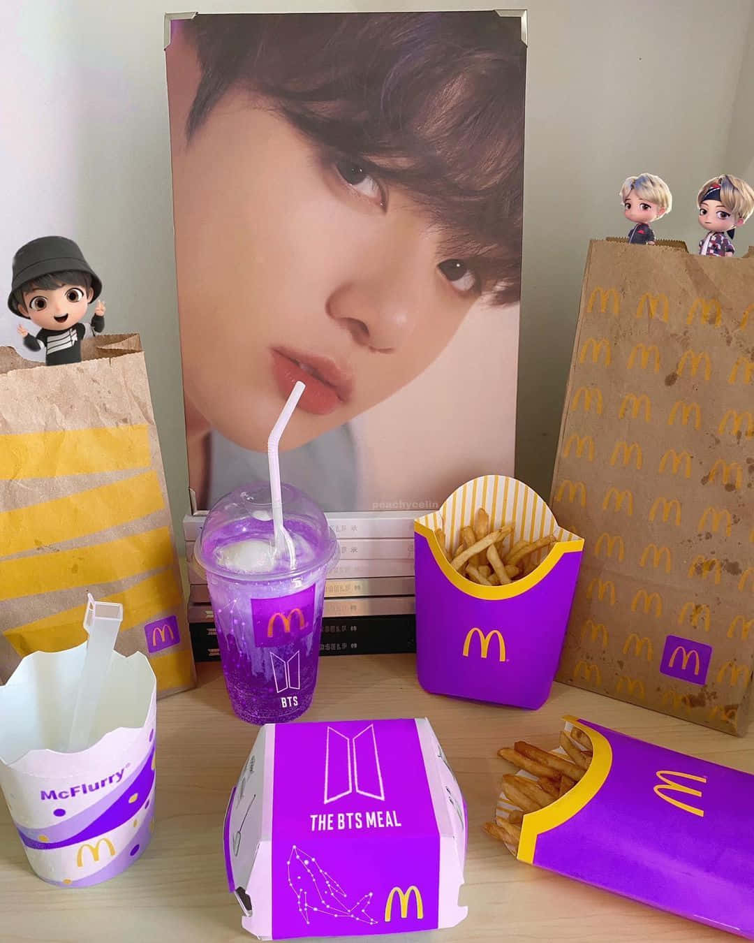 Bts Mac 桌布 壁紙 — 以紫色包裝和一個被小型人偶包圍的成員的大型海報為特色的BTS主題麥當勞餐點。