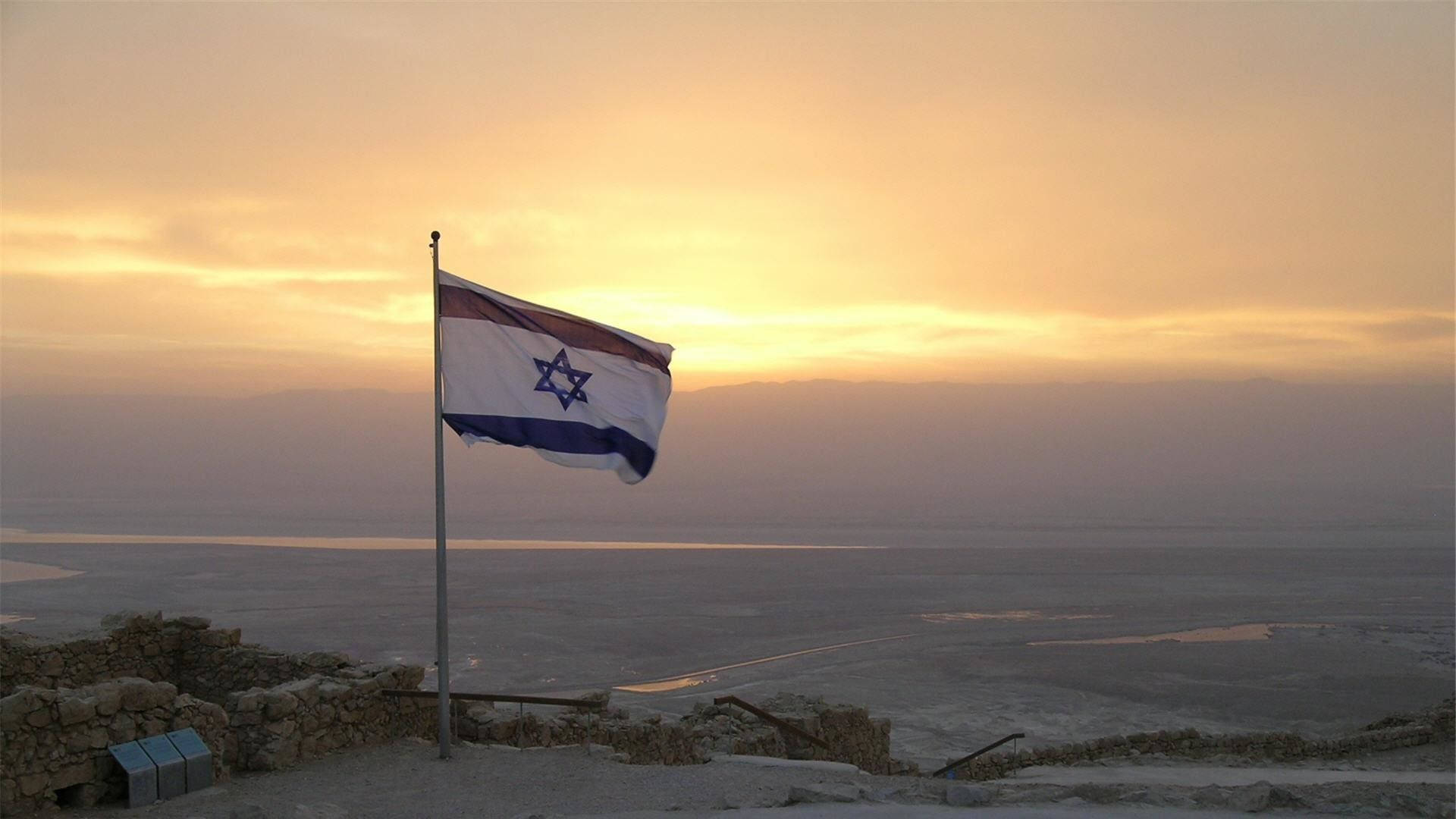 Israel Flag 桌布 壁紙 — 以色列國旗在古老石頭廢墟上自豪地飄揚，俯瞰金色日落下的廣闊鹽灘。