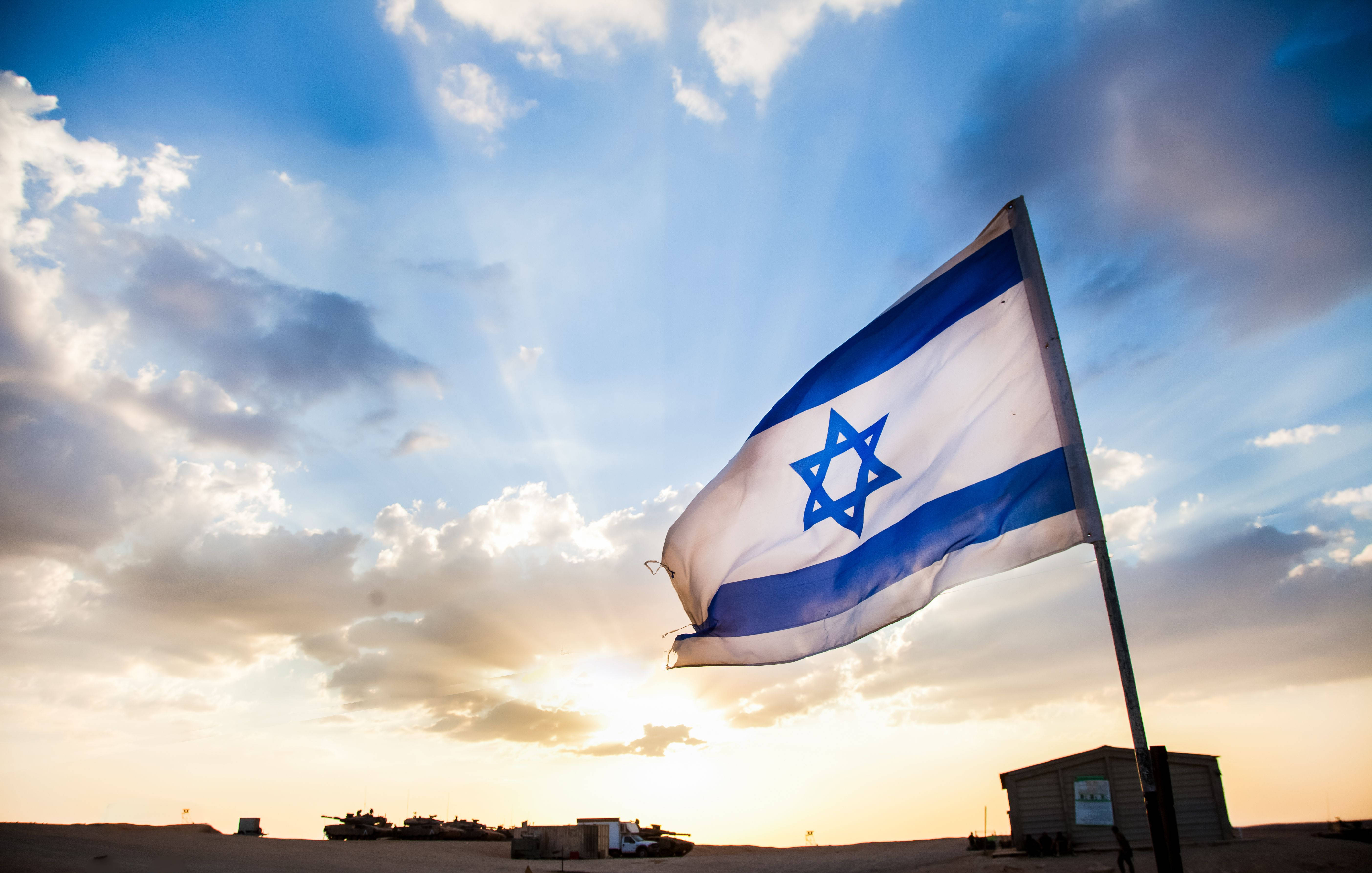 Israel Flag 桌布 壁紙 — 以色列國旗在戲劇性的日落天空下驕傲飄揚，下方沙漠中可見軍用車輛。