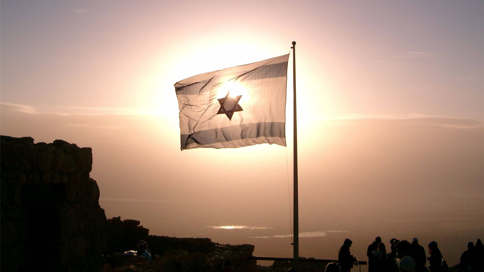 Israel Flag 桌布 壁紙 — 以色列國旗在燦爛的日落天空下自豪地飄揚，下方有剪影人物和岩石地形。