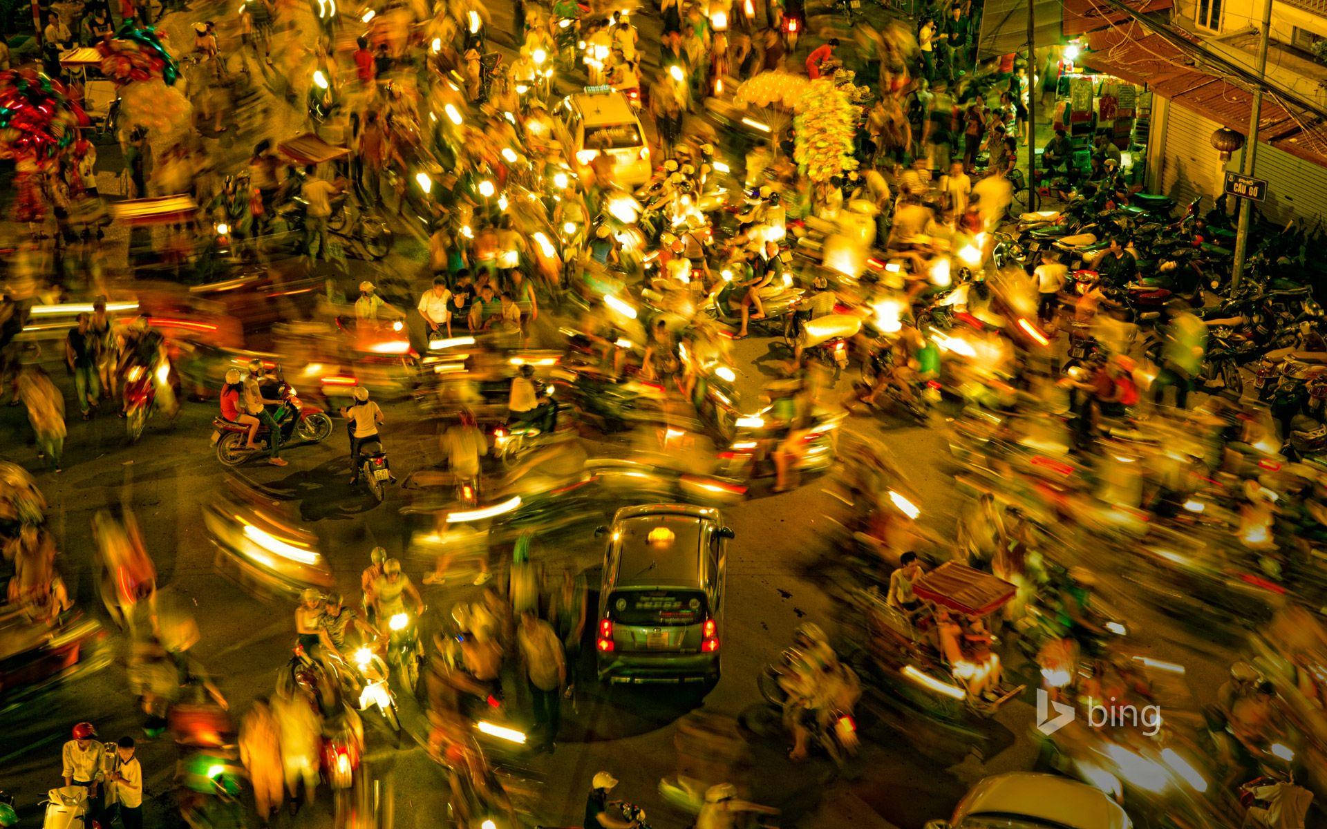Hanoi 桌布 壁紙 — 以長時間曝光捕捉充滿活力的夜街場景，顯示摩托車和行人模糊的動態，並散發溫暖的黃色燈光。
