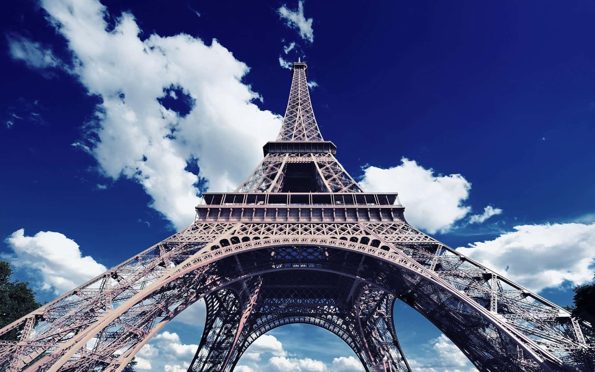 Eiffel Tower 桌布 壁紙 — 以鮮豔的藍天和蓬鬆的白雲為背景，低角度觀看艾菲爾鐵塔複雜的鐵格子結構。