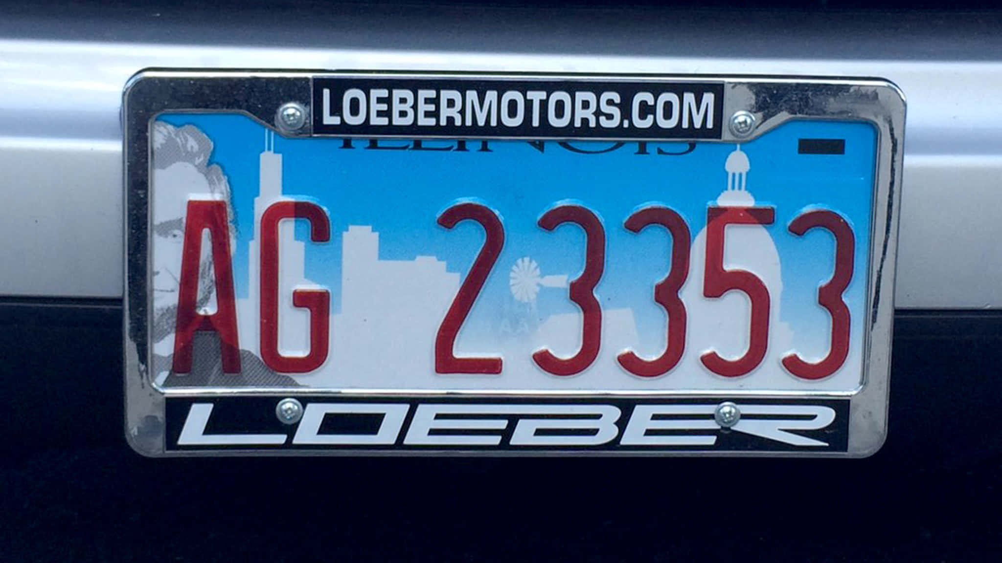 Car License Plate 桌布 壁紙 — 伊利諾州車牌的近距離特寫，紅色字母 AG 23353 鑲嵌在印有 LOEBERMOTORS.COM 的鍍鉻車牌架中。