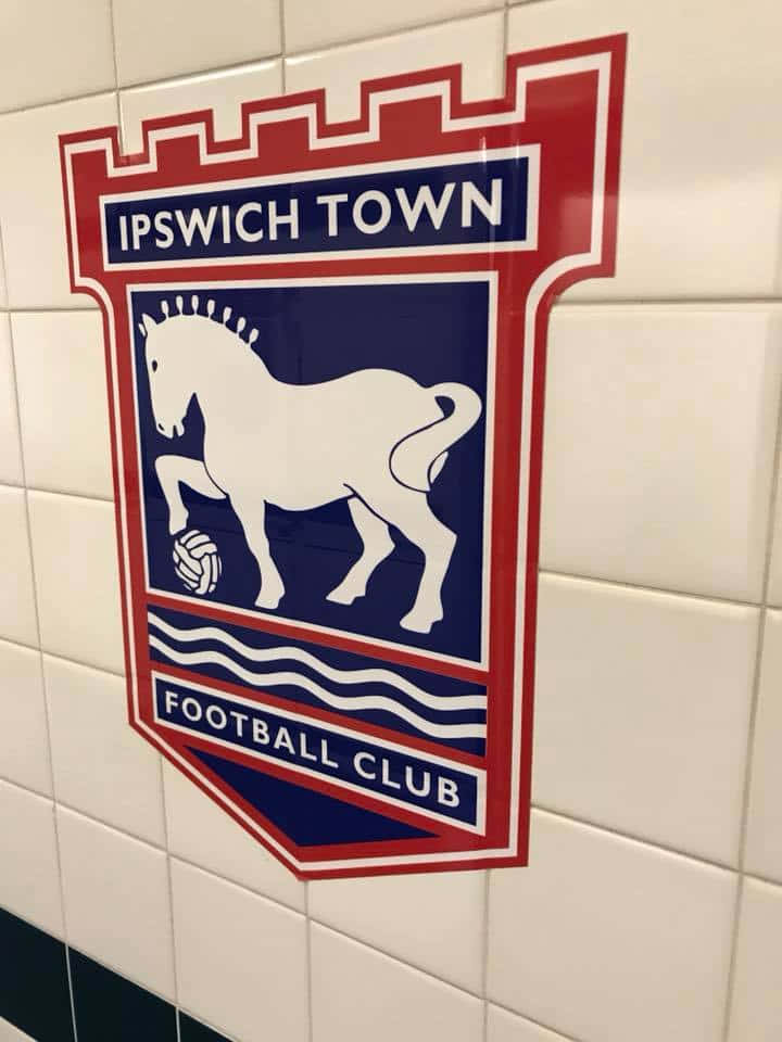 Ipswich Town 桌布 壁紙 — 伊普斯維奇足球俱樂部徽章，上有白馬和足球，藍色背景，安裝在白色瓷磚牆上。