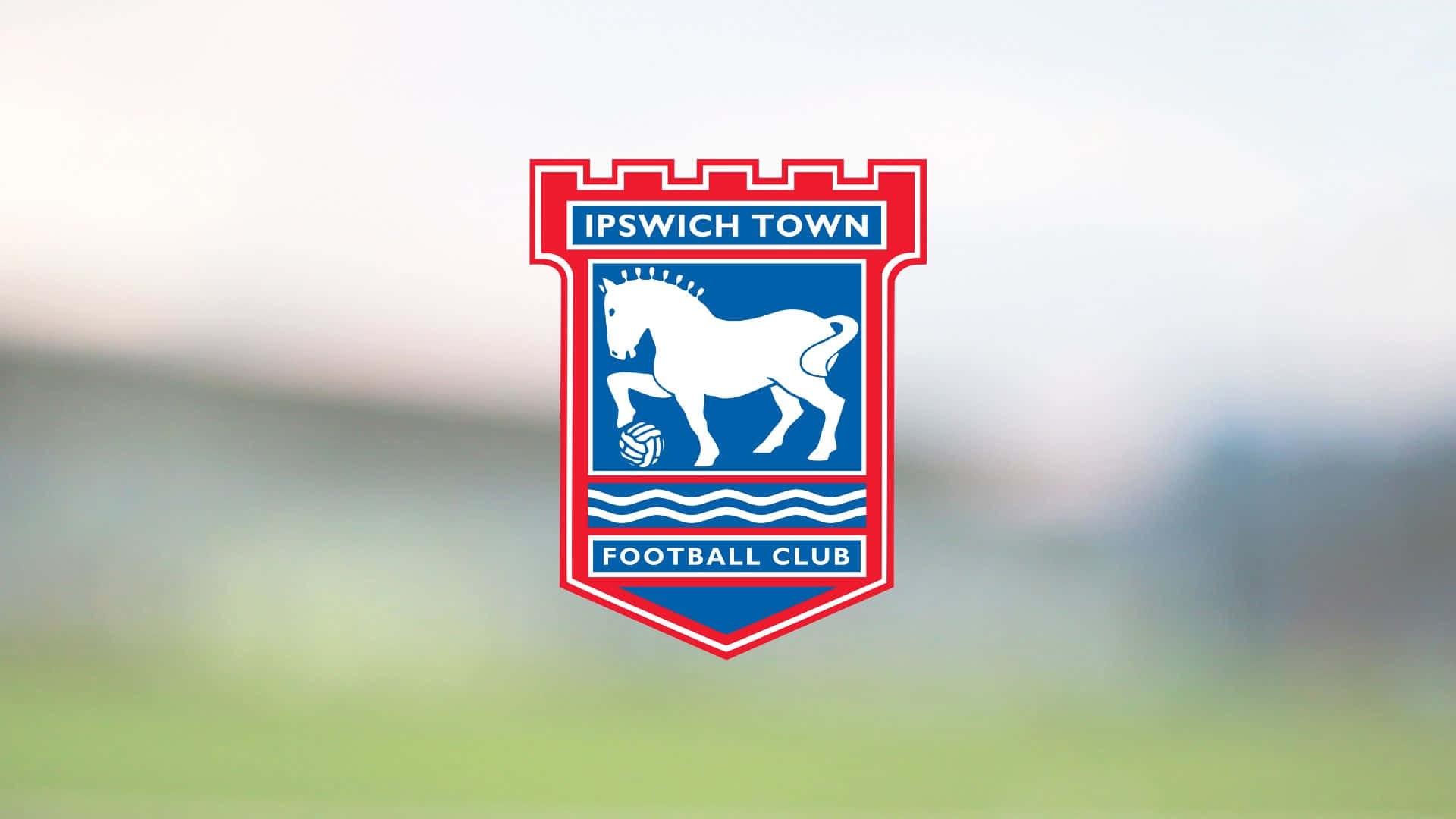 Ipswich Town 桌布 壁紙 — 伊普斯維奇鎮足球俱樂部徽章，藍色盾牌上有白馬和足球，背景為模糊的綠色球場。