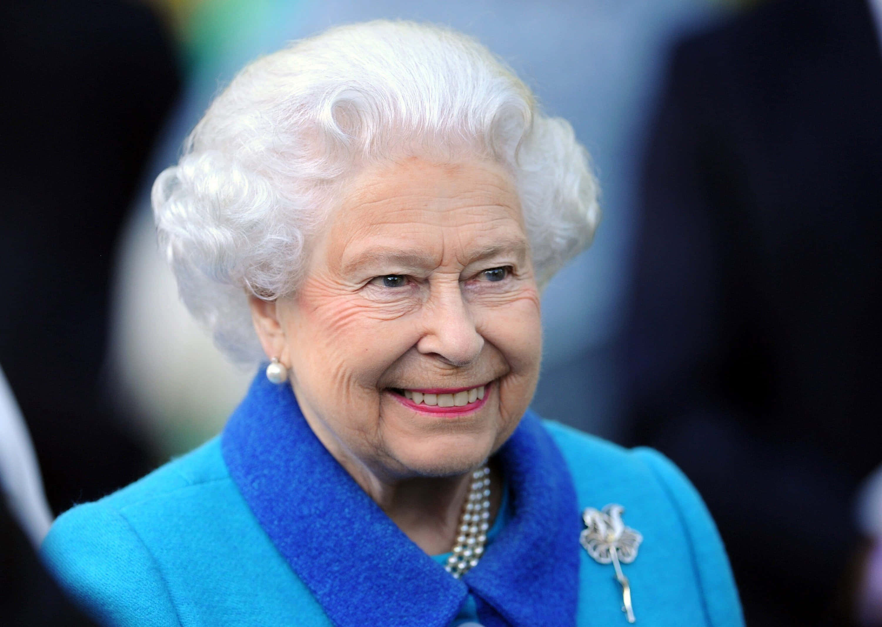 Queen Elizabeth 桌布 壁紙 — 伊莉莎白二世女王的特寫肖像，她穿著飾有珍珠和胸針的鮮藍色外套，溫暖地微笑。