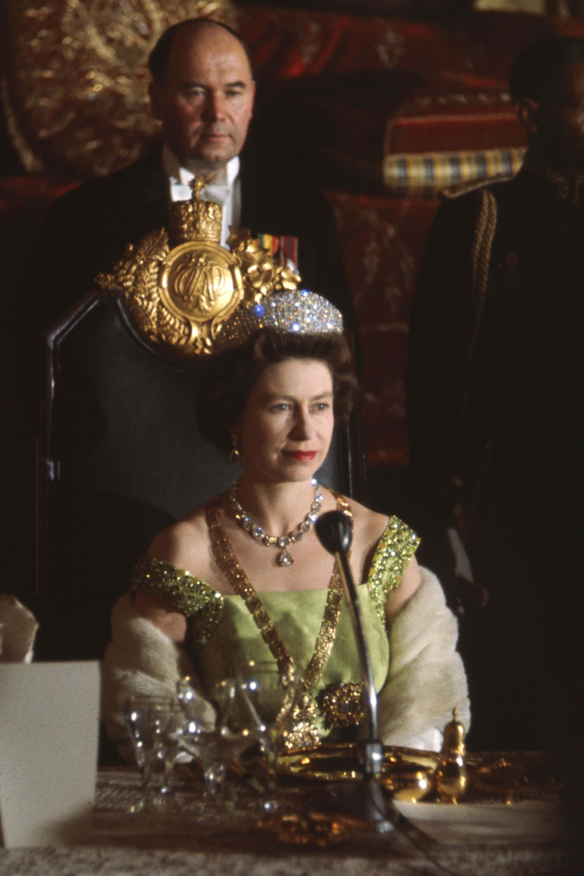 Queen Elizabeth 桌布 壁紙 — 伊莉莎白二世女王身穿綠色長袍和鑽石头飾，端莊地坐在正式活動中，身後椅子上放著一頂金色王冠。
