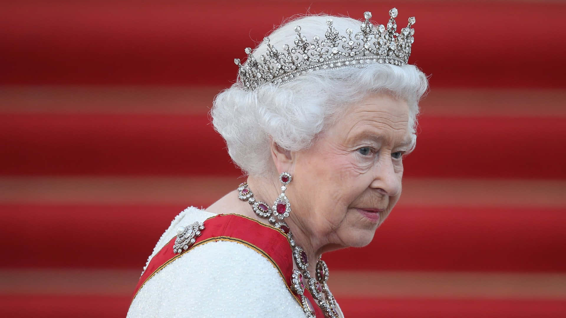Queen Elizabeth 桌布 壁紙 — 伊莉莎白二世女王身穿華麗服飾，佩戴閃爍鑽石冠冕和紅寶石耳環，背景是鮮豔的紅地毯。
