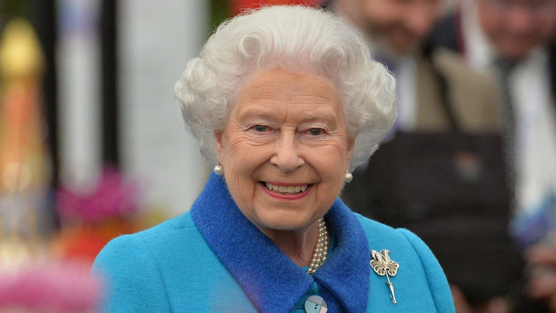 Queen Elizabeth 桌布 壁紙 — 伊莉莎白二世女王身穿鮮藍色外套，佩戴珍珠首飾和三葉草胸針，溫暖地微笑。