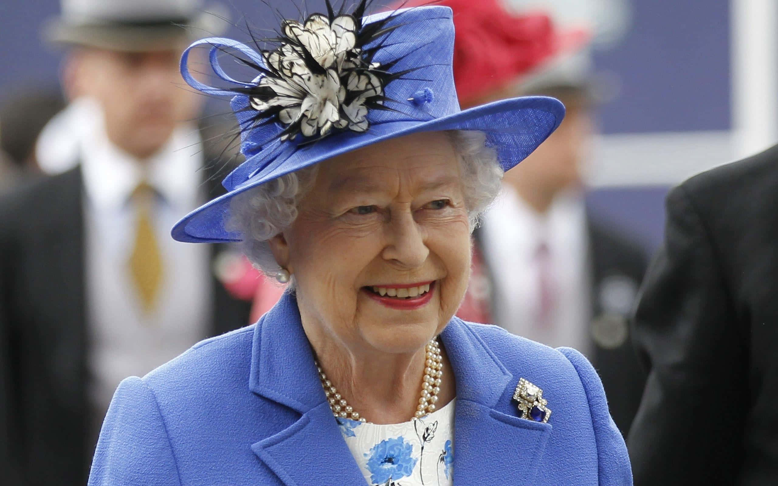 Queen Elizabeth 桌布 壁紙 — 伊莉莎白二世女王身穿鮮藍色外套和搭配的帽子，帽上飾有黑白羽毛，溫暖地微笑。