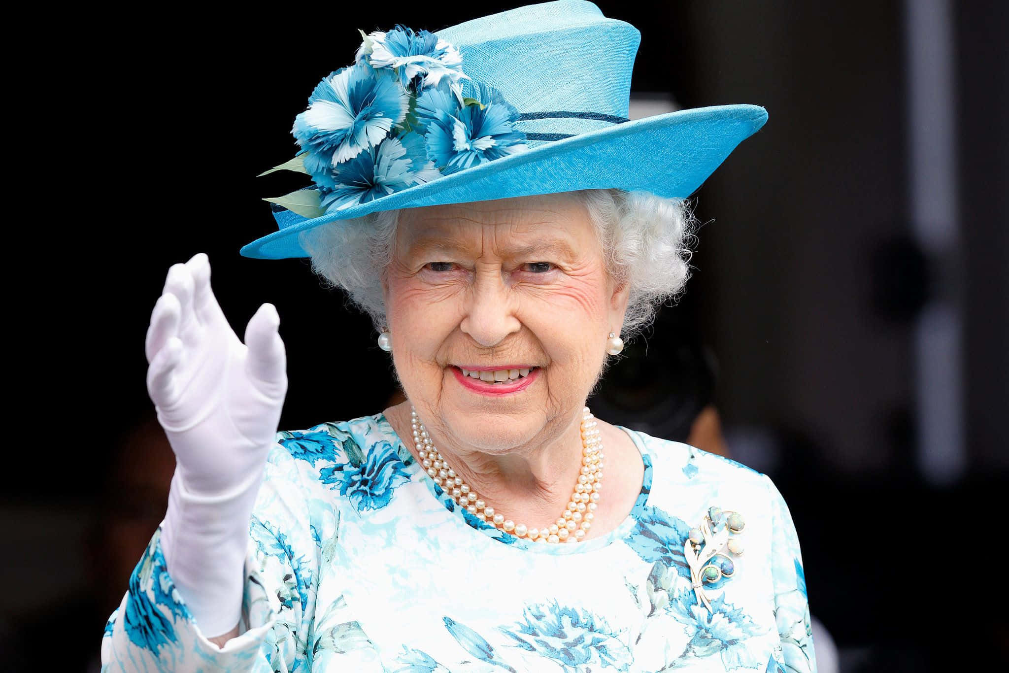 Queen Elizabeth 桌布 壁紙 — 伊莉莎白二世女王身穿鮮豔的藍色花卉套裝，頭戴飾有藍色花朵的配套帽子，溫暖地微笑揮手。