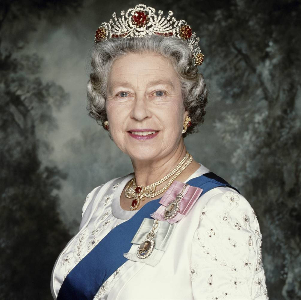 Queen Elizabeth 桌布 壁紙 — 伊莉莎白二世女王面帶微笑，威嚴地佩戴著鑲有寶石的王冠、珍珠和藍色肩帶，背景是柔和的森林。