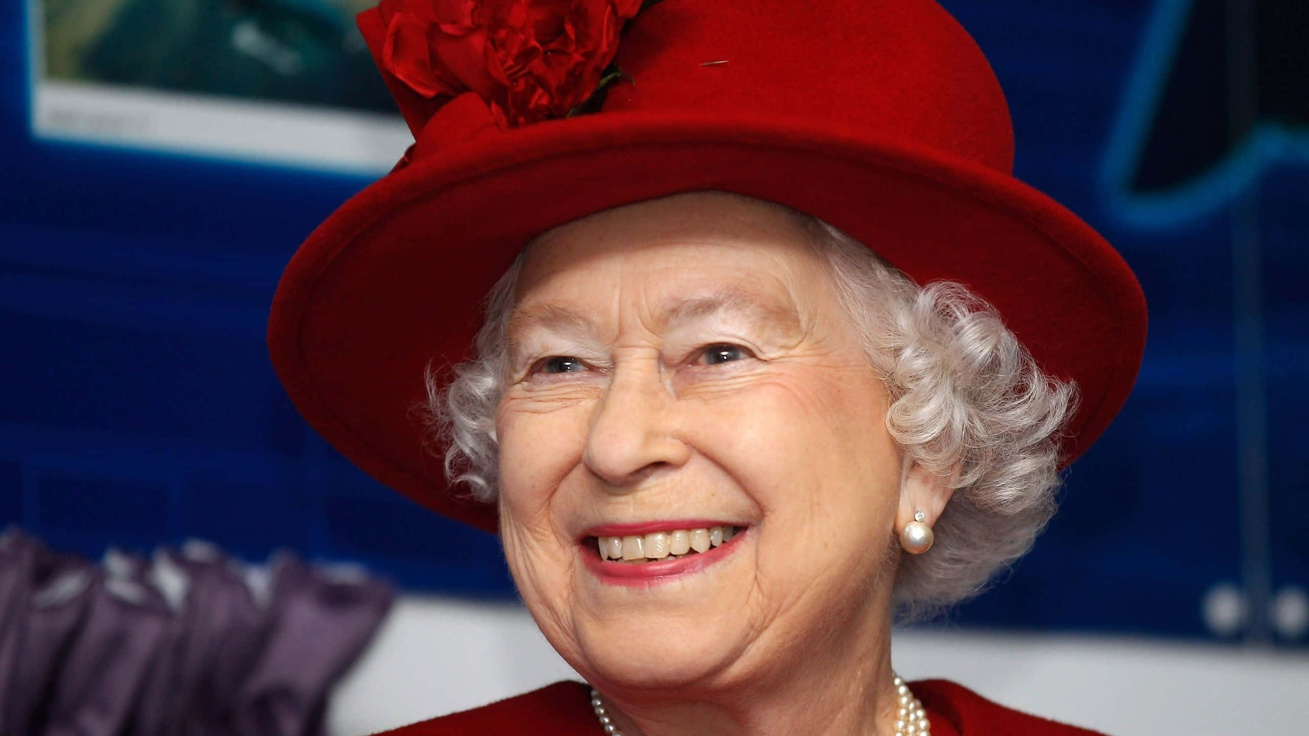 Queen Elizabeth 桌布 壁紙 — 伊莉莎白二世女王面帶溫暖微笑，戴著一頂飾有大紅花的搶眼紅帽，搭配紅色套裝。