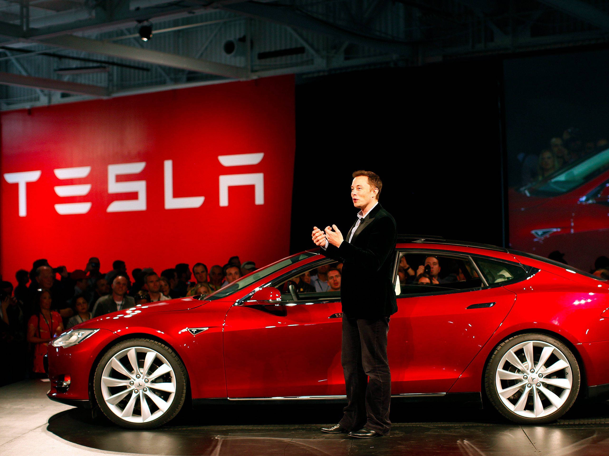 Elon 桌布 壁紙 — 伊隆·馬斯克在明亮的活動燈光下，在舞台上向人群展示一輛光亮的紅色Tesla Model S。