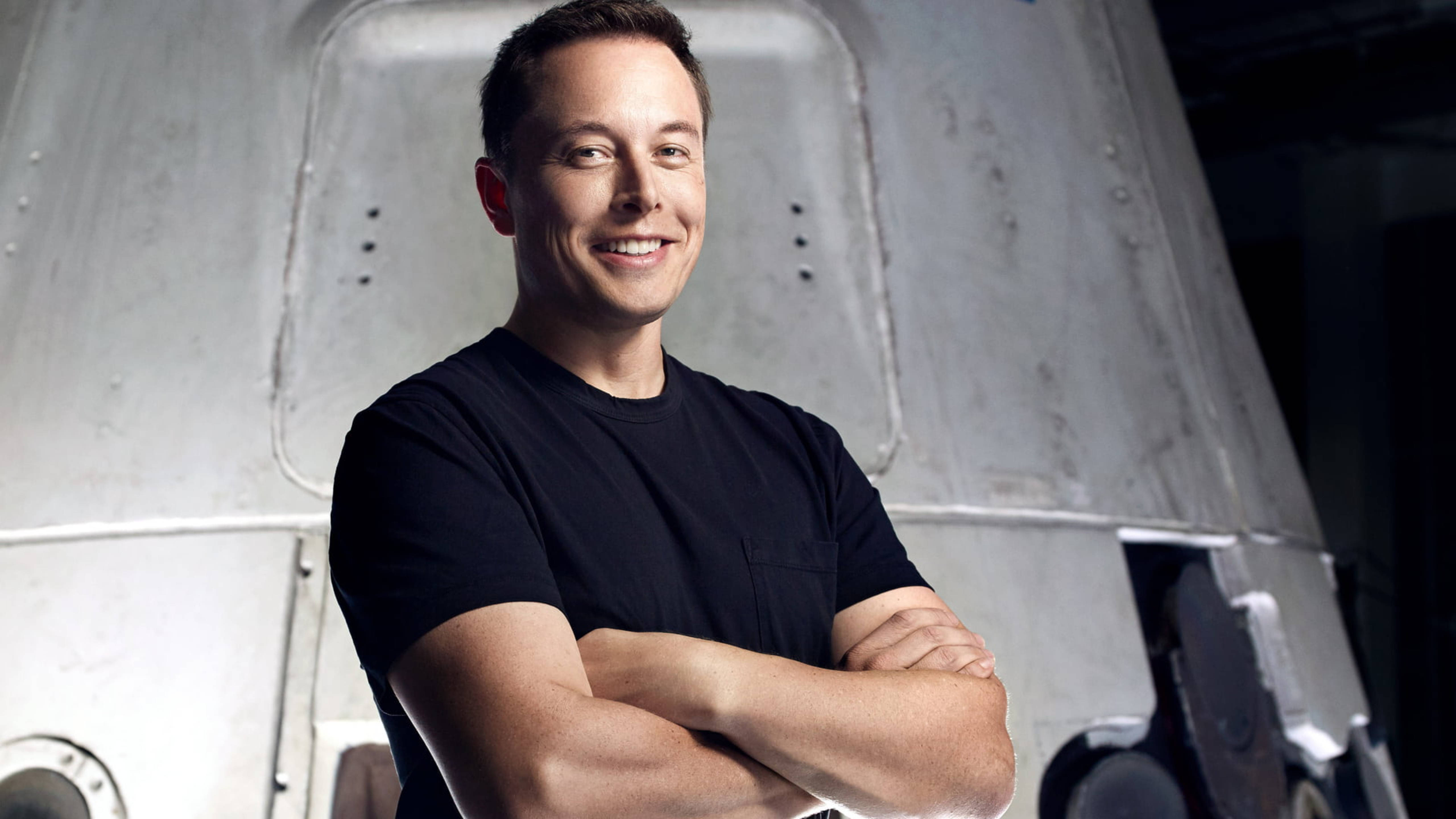 Elon 桌布 壁紙 — 伊隆·馬斯克在燈光昏暗的機庫中，雙臂交叉，自信地微笑，身後是一艘大型金屬太空船。